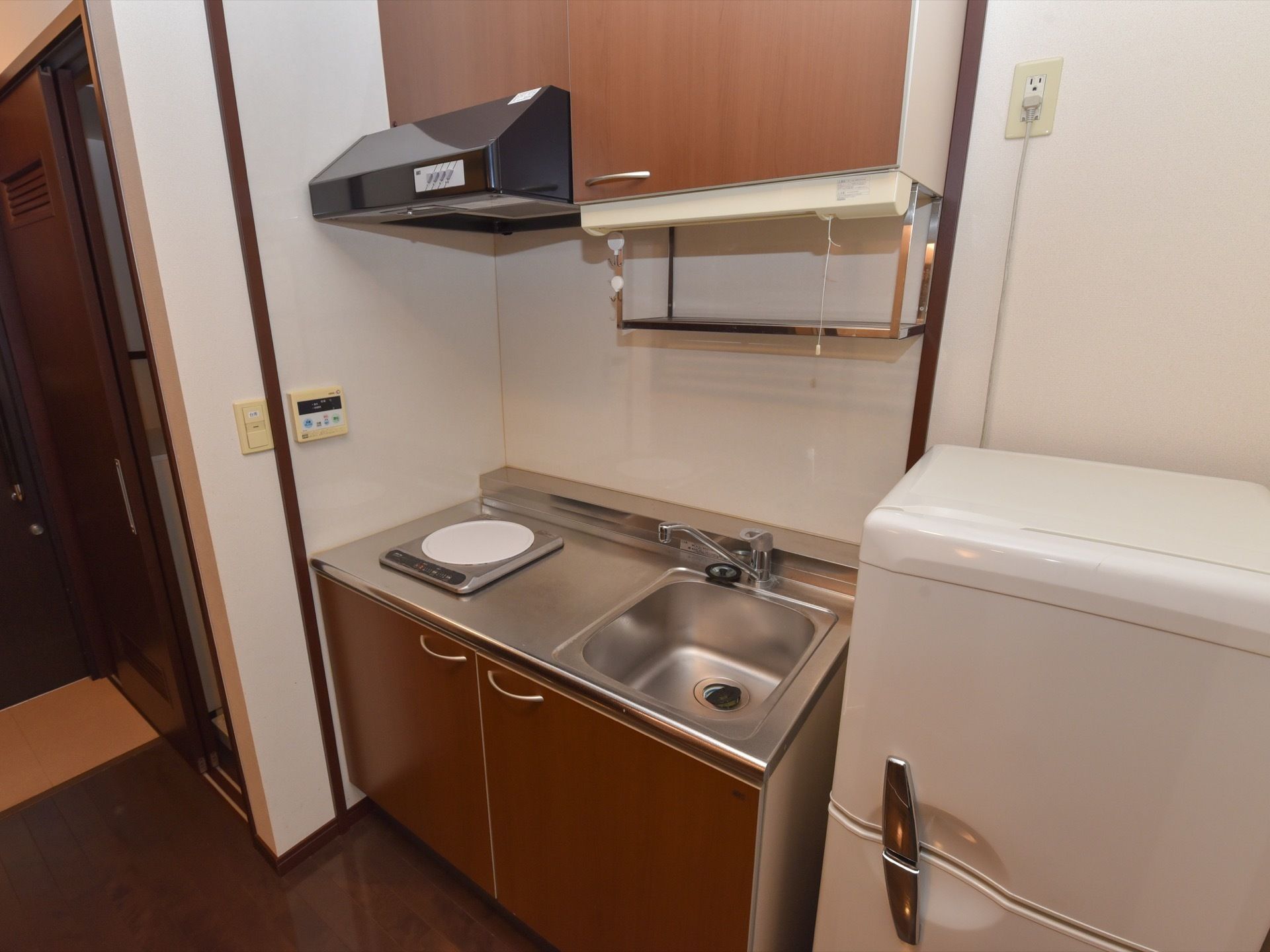 ミニキッチンがと冷蔵庫がある。キッチン左側に電気コンロが１台あり、上に換気扇がある。右側にシンクがあり、上に収納棚が付いている。