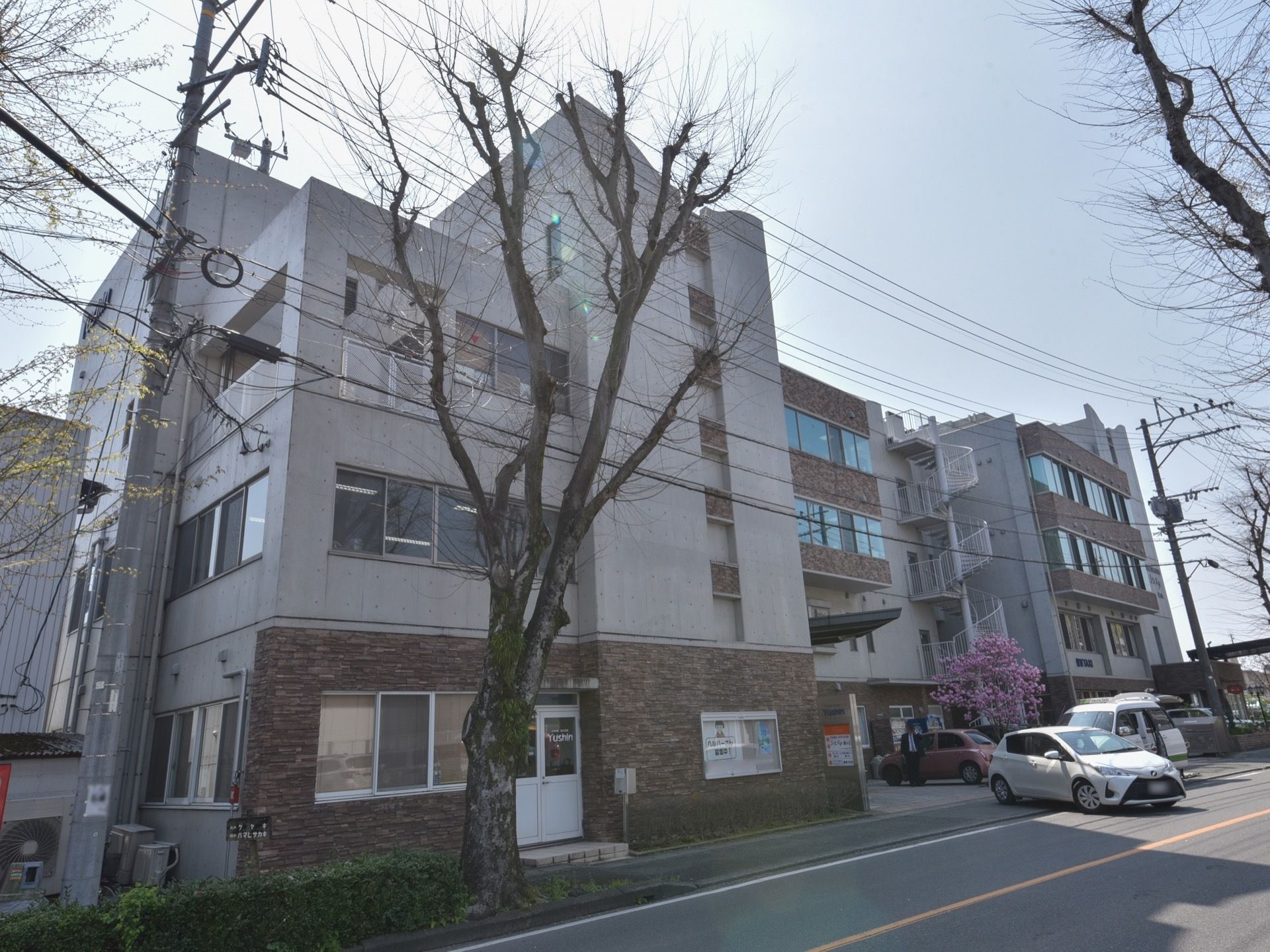 広い道路に面したグループホームで、建物の前に駐車スペースがある。白いらせん状の階段が設置されている。