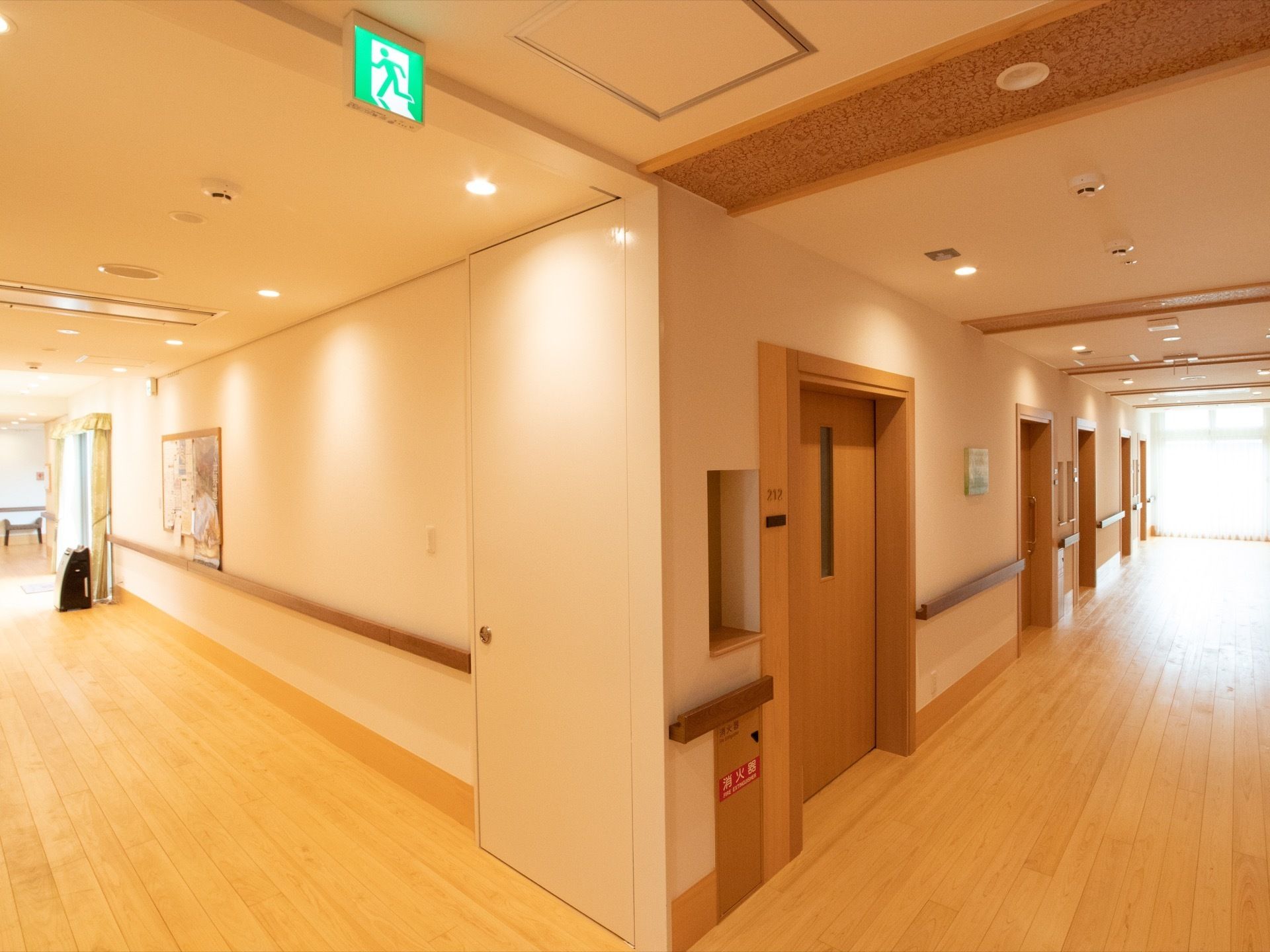 廊下の角の様子で、床はフローリングになっている。防火扉が設置されており、天井には避難誘導灯がある。壁には絵画が飾られている。