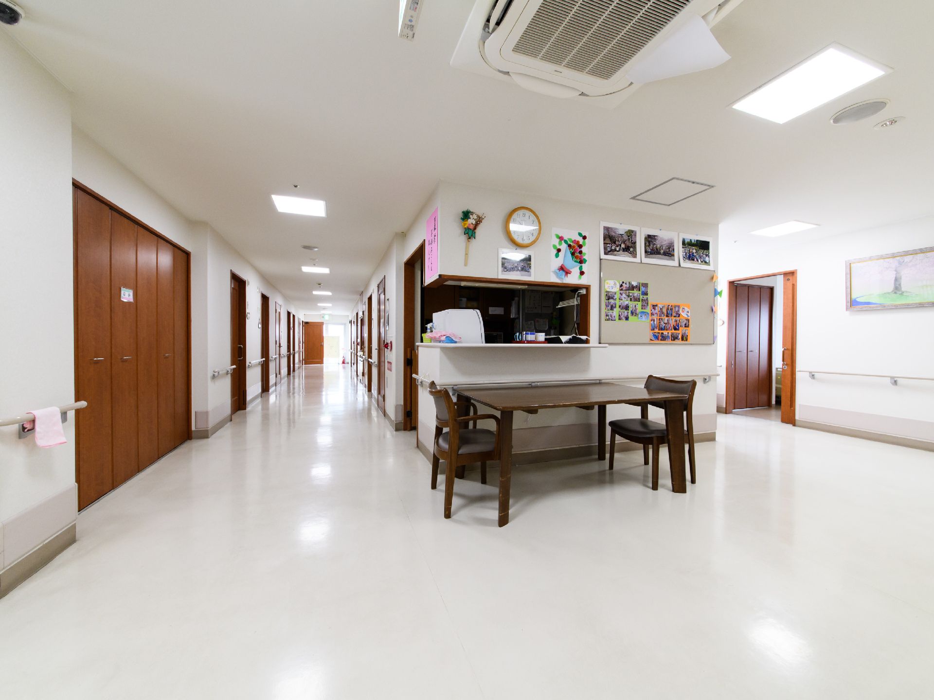 観音開きのドアが廊下の左手に見える。手すりが壁の左右に取り付けてある。キッチンが右手に見える。手前にテーブルと椅子が置いてある。