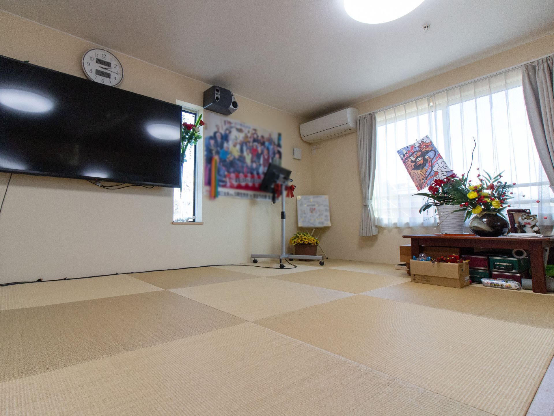 正方形の畳マットを敷き詰めた共有スペースがある。壁に大型液晶テレビが取りつけられている。窓際に棚が置かれ、花などが飾られている。
