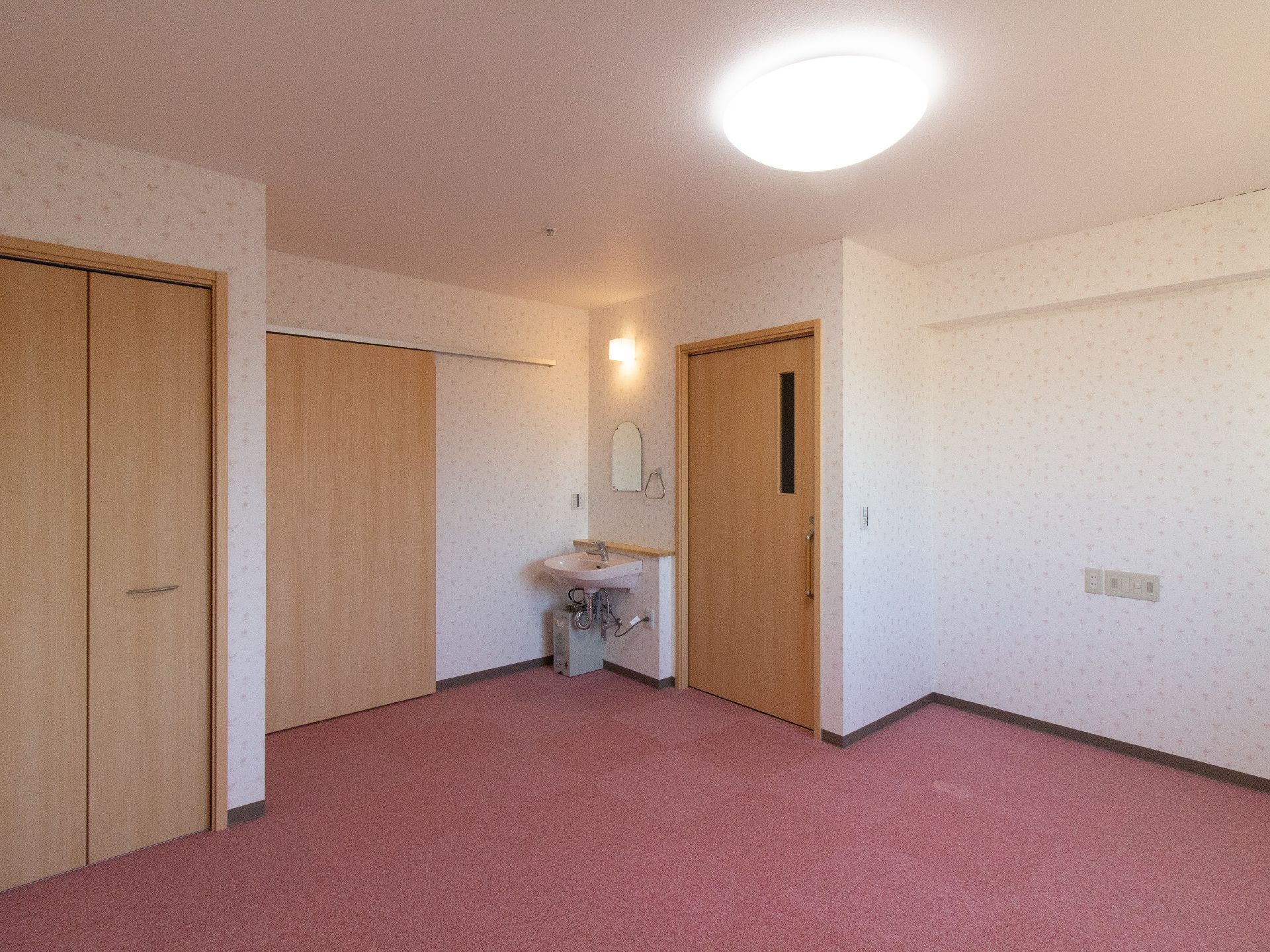 ピンクの絨毯が敷かれた居室で、入口横に洗面台があり、その隣にトイレへのドアがある。左手前にクローゼットがある。