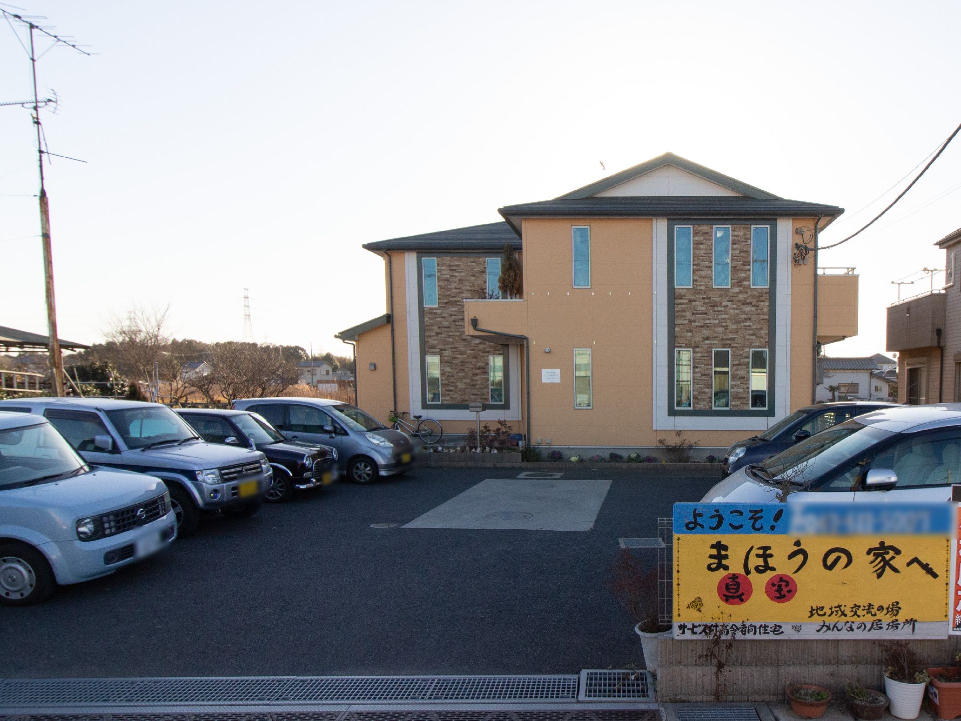 駐車場の入口にはペンキで手書きした看板が立っている。２階建てのホームは、濃い茶色のタイルと薄茶色の外壁からなる。