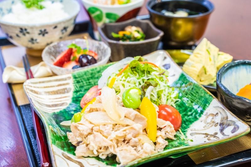 松花堂弁当の箱に扇型に抜いて胡麻を散らしたご飯、天ぷらと野菜の炊き合わせなどが盛り付けられている。果物の小鉢とデザートも並んでいる。