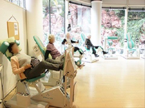 白木の床の上に、筋肉トレーニングマシーンが６台並んでいる。大きな窓ガラスの外には庭の緑が見える。４名の入居者様が汗を流している。