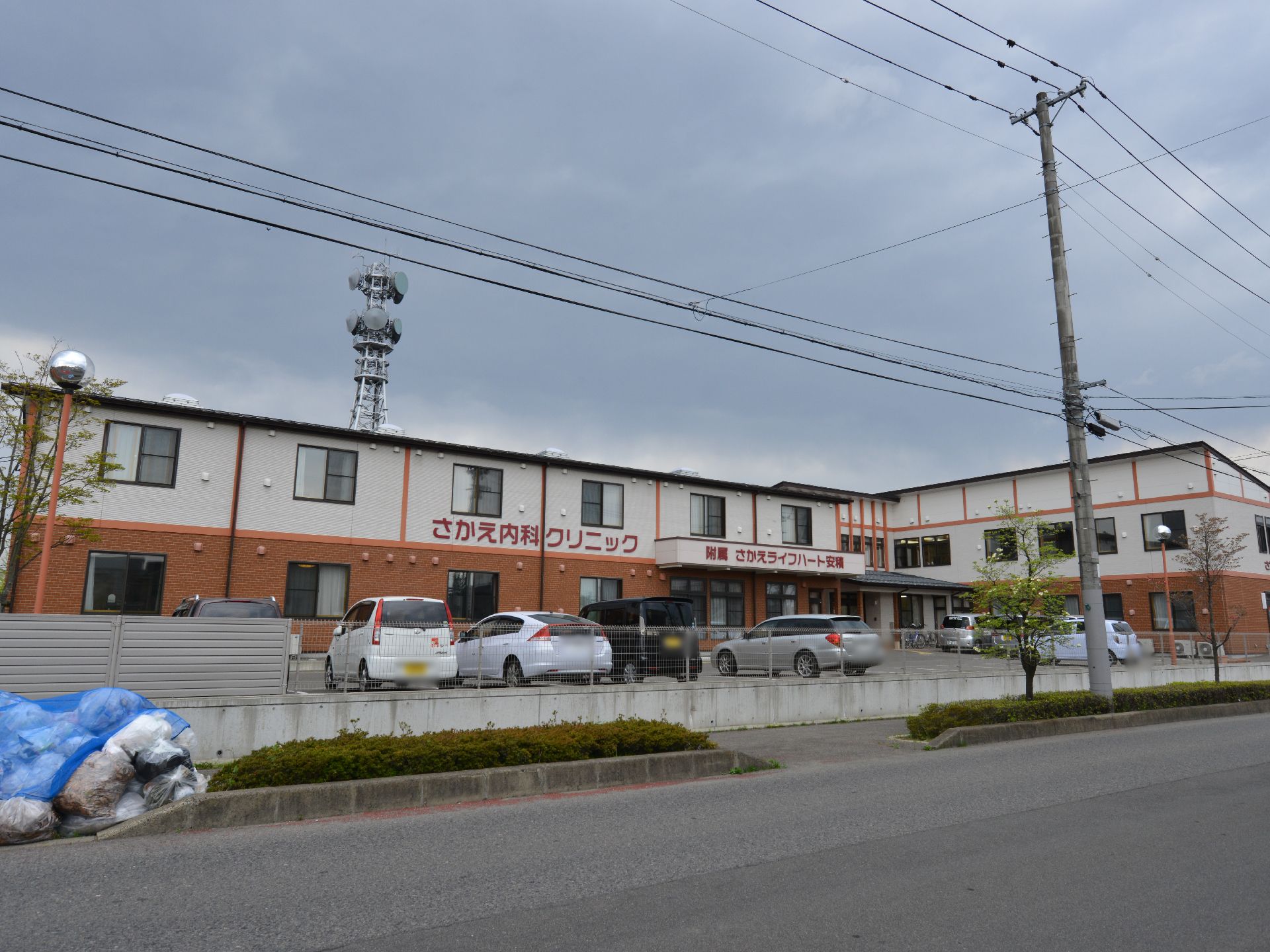 施設外観。建物の色は白と茶系で落ち着いている。広い駐車場があり、となりにはさかえ内科クリニックがある。