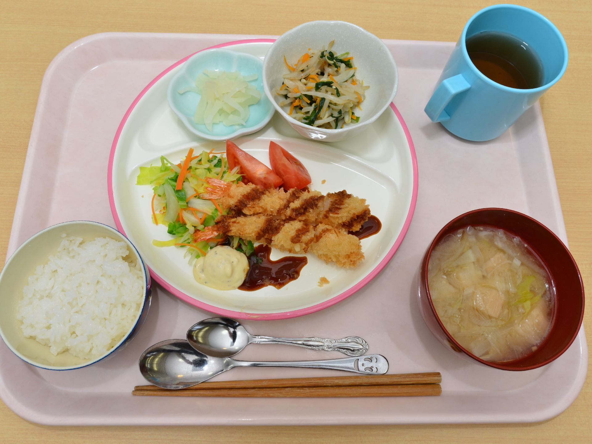 施設利用者の食事の一例である。主食、汁物、主菜、副菜、お漬物、お茶がプレートに乗っている。お箸と大小ふたつのスプーンが用意されている。