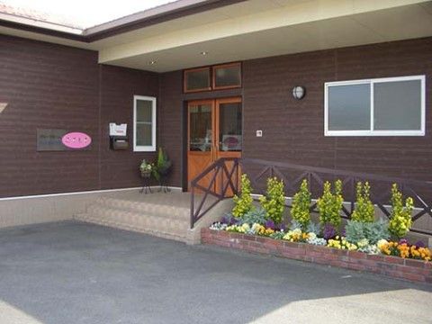 建物の壁はこげ茶色で、玄関ポーチの部分はベージュ色の屋根が付いている。周囲をきれいな花々や草木で囲まれた玄関である。