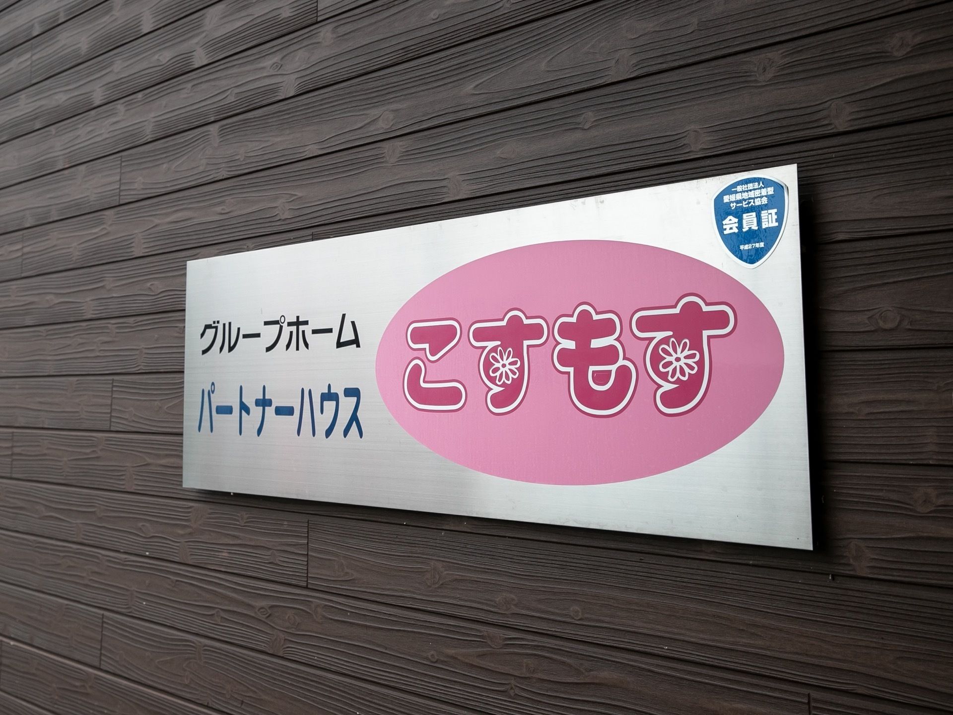 銀色のホームの看板が、木目調の壁のタイルに設置してある。名称はピンクの楕円に囲まれており、花も描かれている。