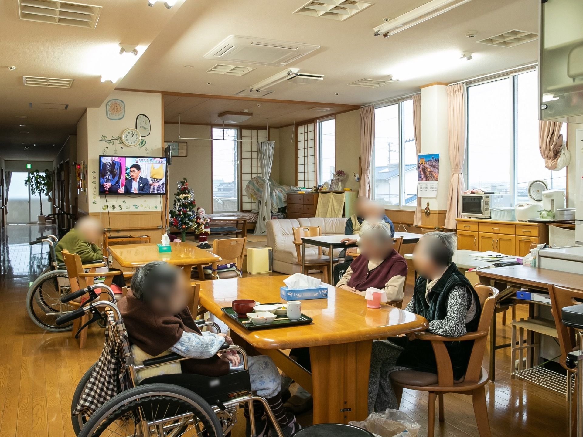 ４人がけのテーブルには、肘掛のある椅子が設置してある。入居者様は椅子か車椅子に座って、楽な姿勢でテレビを観て過ごしている。