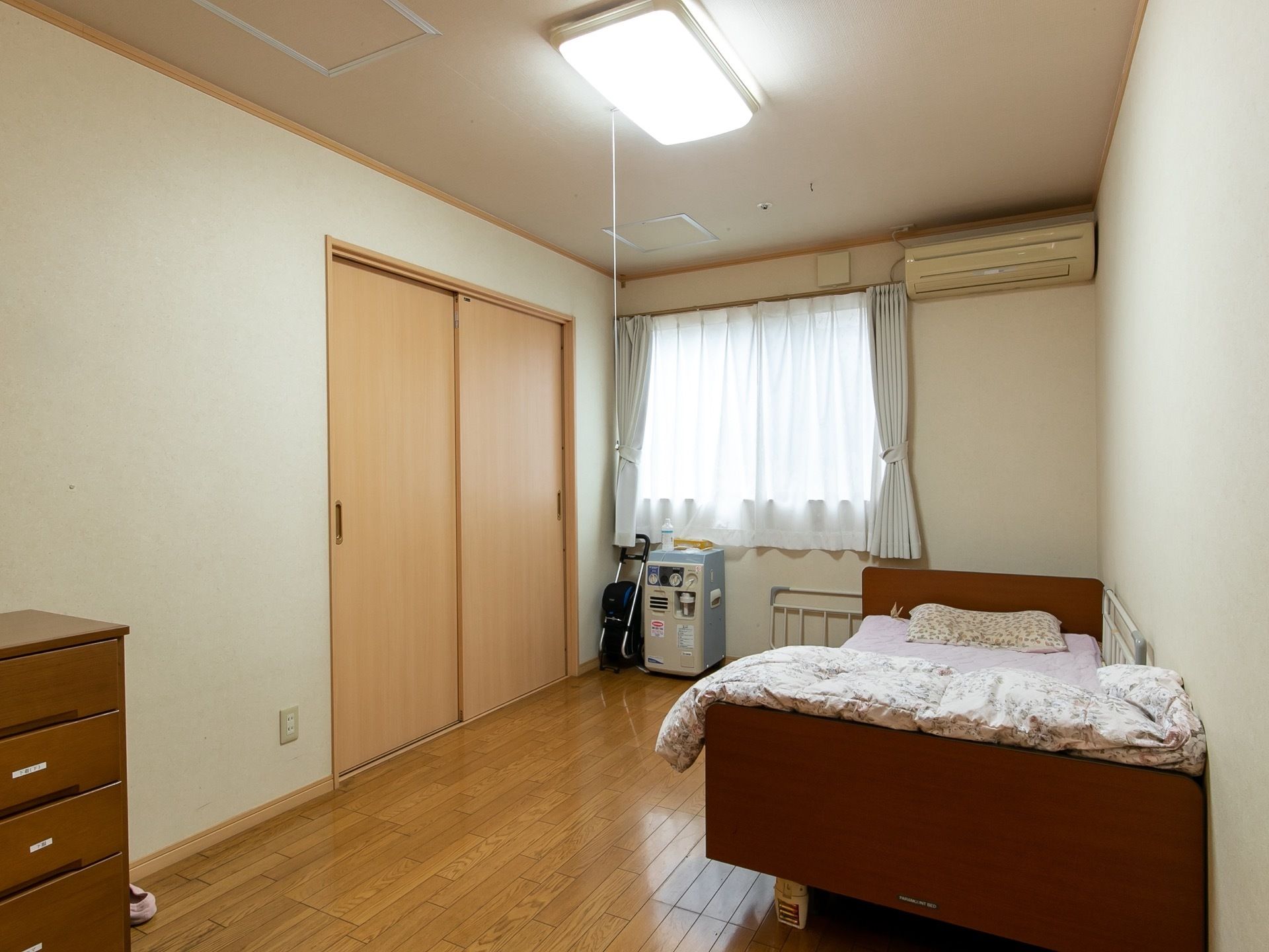 窓から日射しの差し込む明るい居室である。セミダブルの介護用ベッドやエアコン、LEDライト、収納スペースが完備されている。
