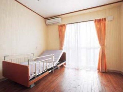 リモコン付きのリクライニングベッドが置かれた居室である。ワンルームの部屋でエアコンが設置されている。