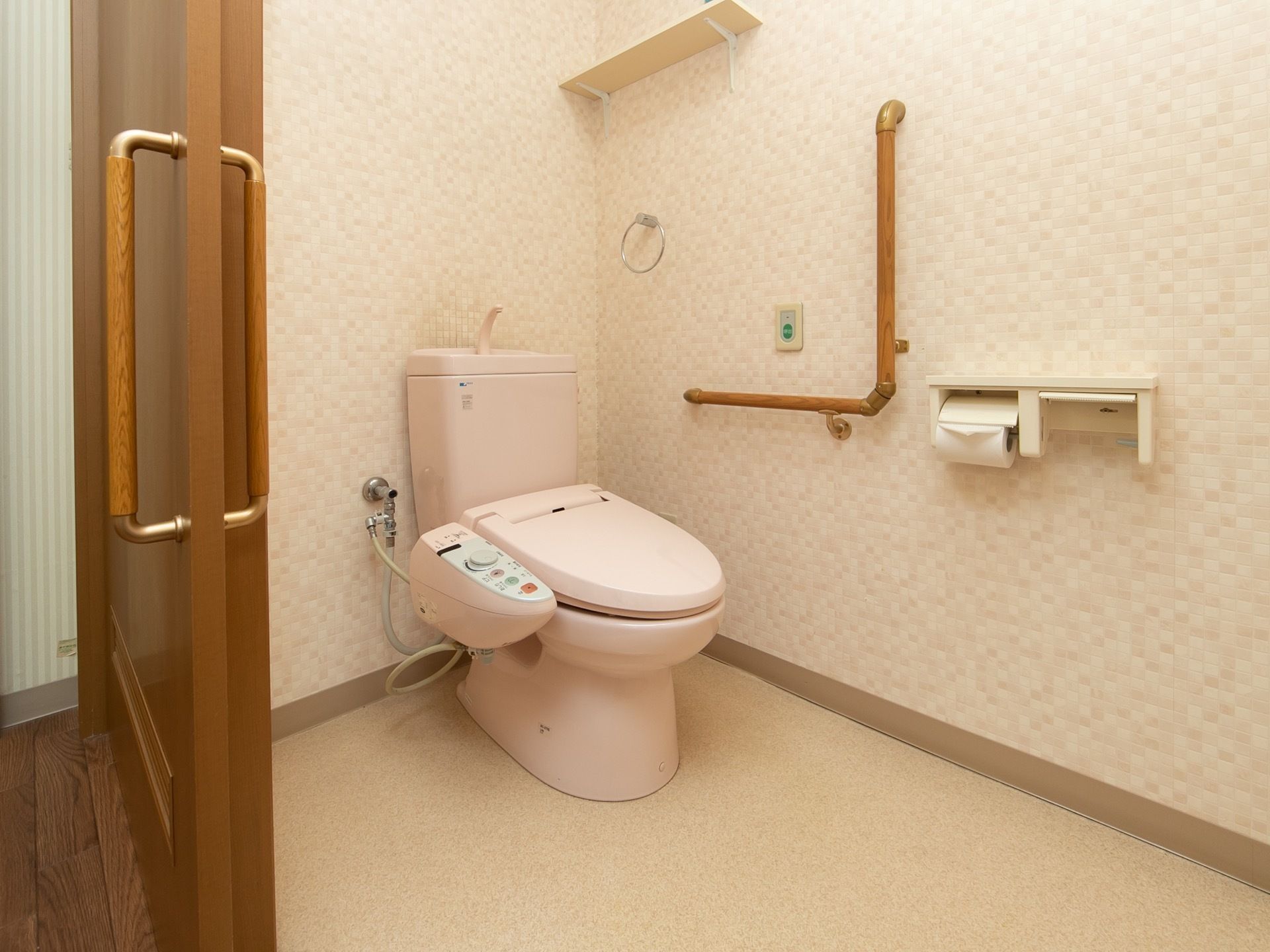 居室内にあるピンク色のトイレである。便器のそばの壁にはＬ字型の手すりや、タオル掛けが設置されている。