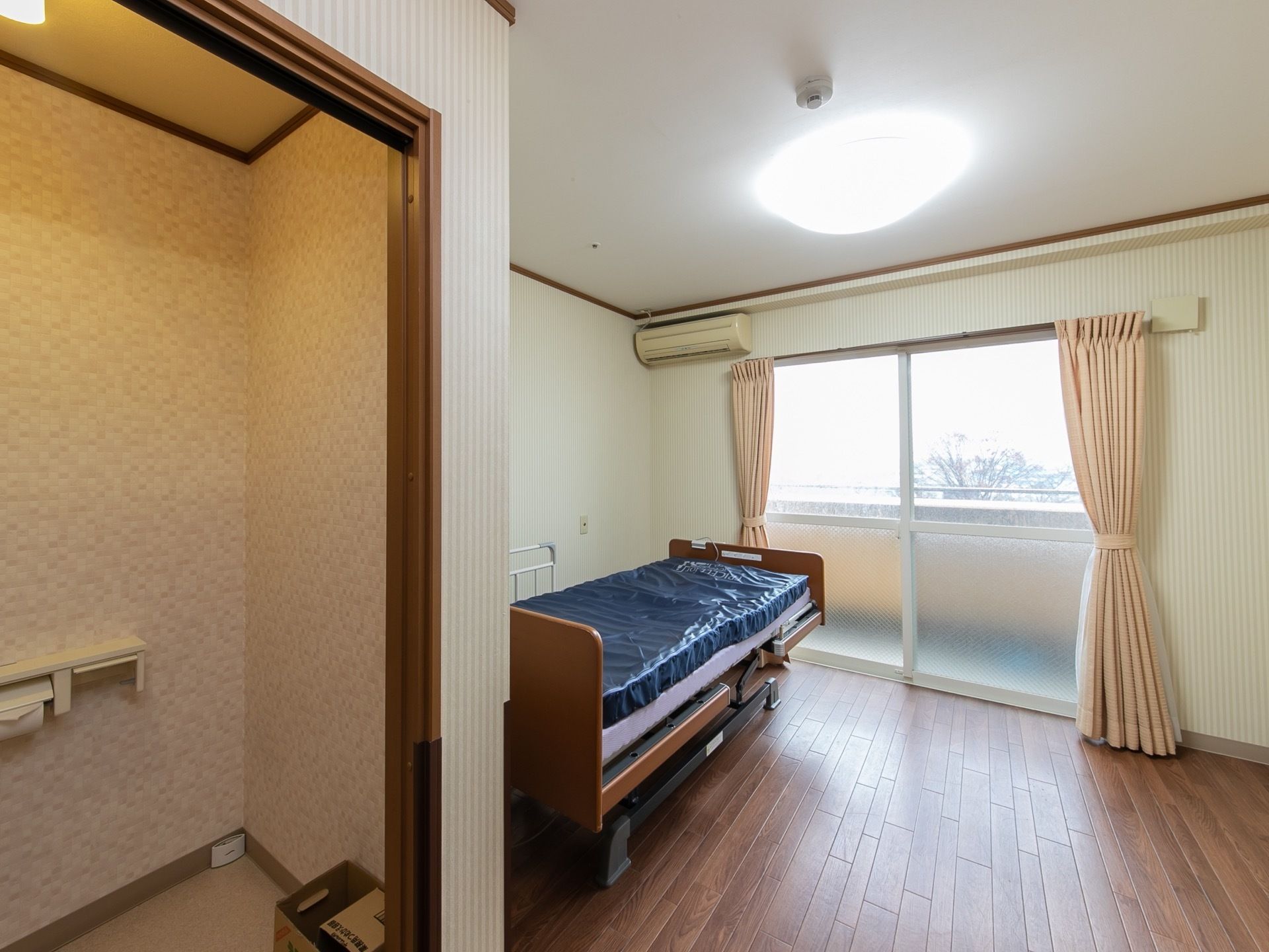 ベランダ付きの日当たりの良い居室である。窓辺には幅が広く、リクライニング可能なベッドが備わっている。