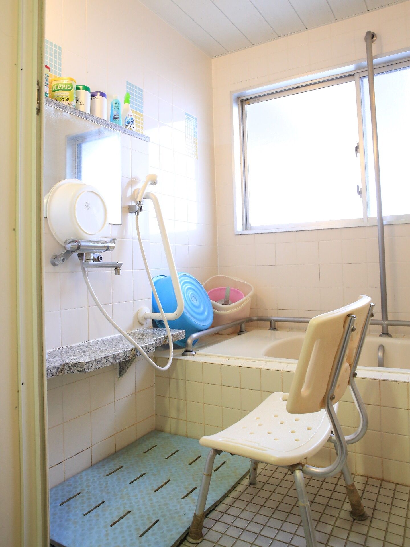 浴室には大きなすりガラスの窓がある。入浴剤や掃除道具などが整理されて置かれている。背もたれのついた椅子がある。