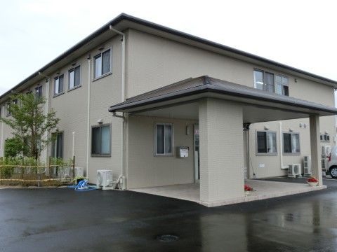 箱型の２階建てで、壁は落ち着いたオフホワイト。玄関周りはアスファルトで舗装されており、ひさしの下にはプランターが置かれている。