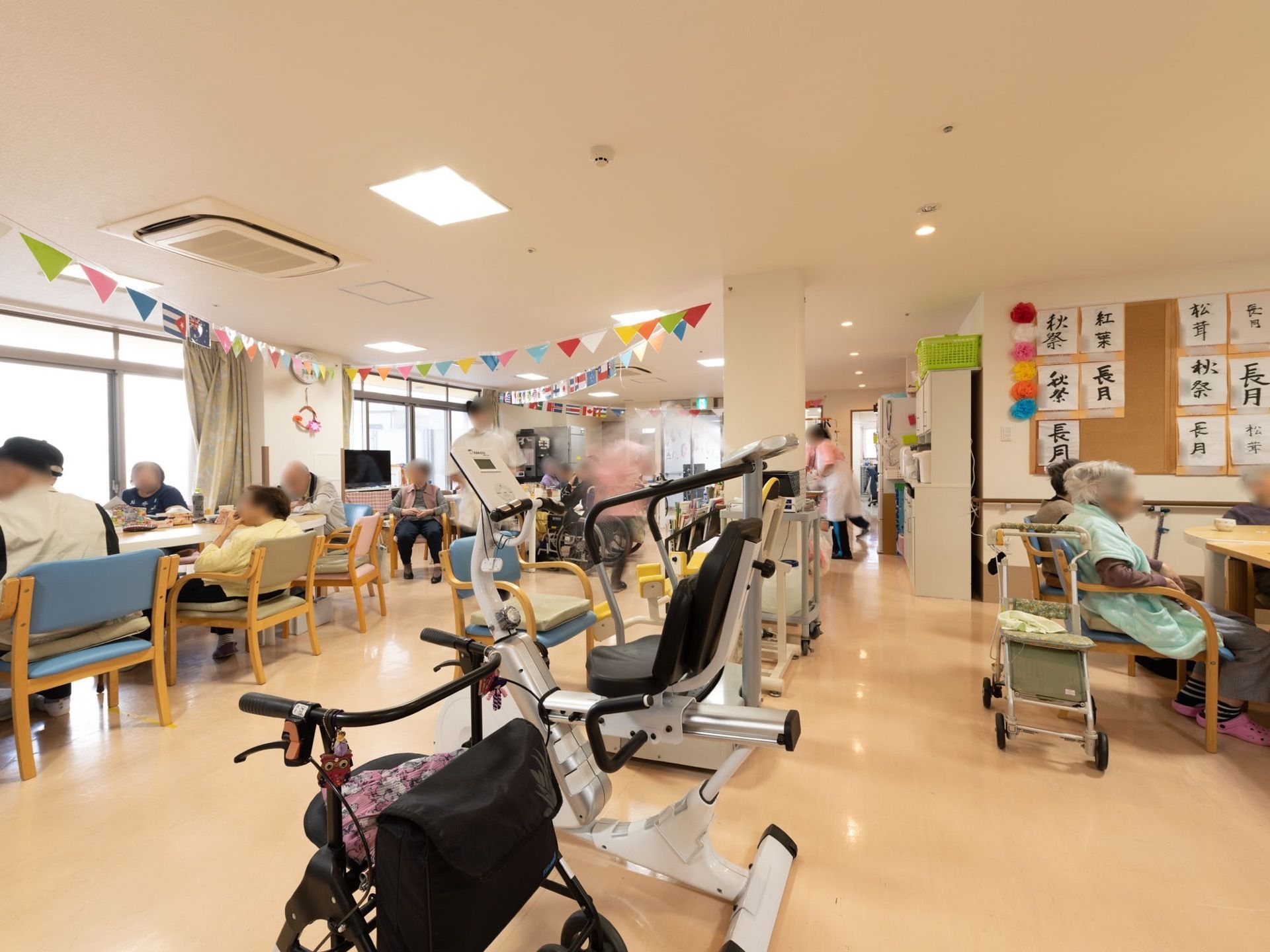 共有ペースの壁や天井はレイアウトされ、スタッフの姿やテーブルセットの椅子に入居者様、皆さまが腰掛けられている姿が見える。