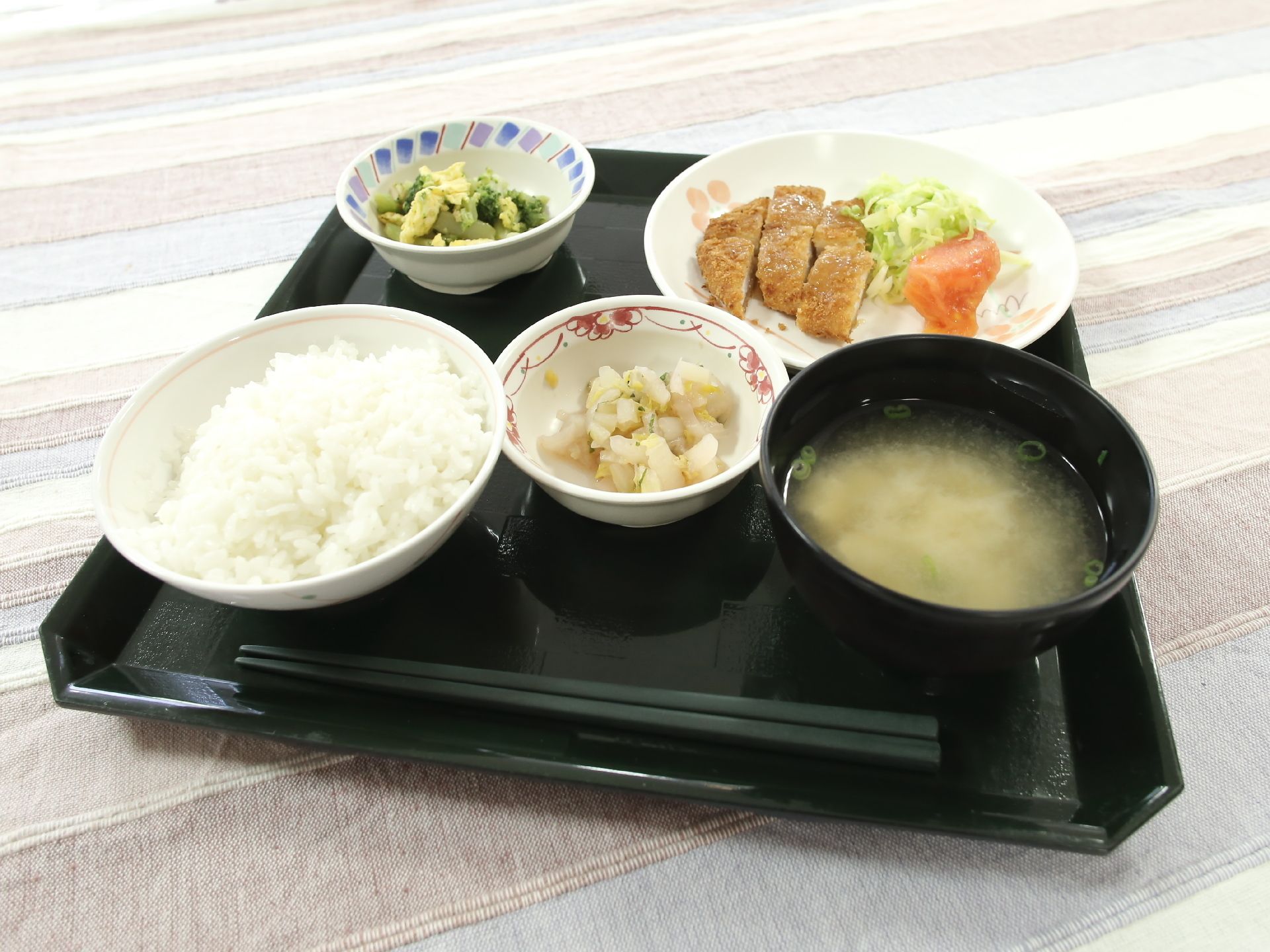 黒いお盆の上に様々な食べ物が置かれている。左側にはご飯があり、右側には味噌汁がある。中央と左上には小鉢がある。右上にはメインのおかずがある。