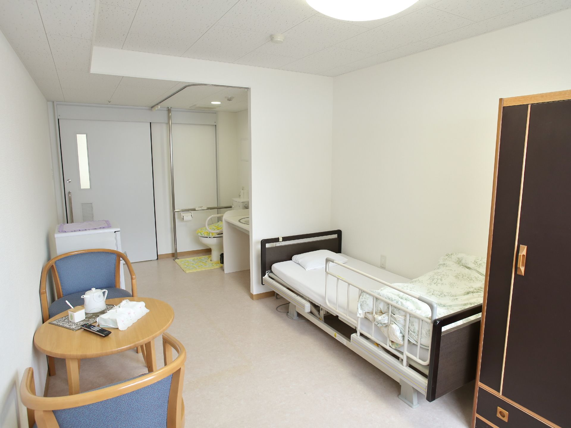 入ってすぐの所に、トイレと洗面スペースが用意されている。介護ベッドには柵が取りつけられている。カフェテーブルと椅子、タンスが置かれている。