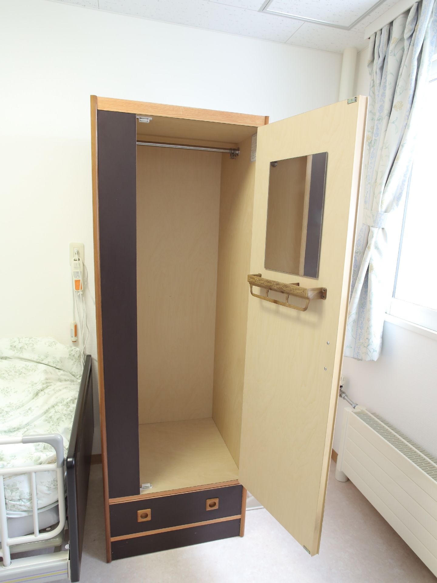 高さのある茶色のクローゼットが置かれている。衣類が掛けられるよう、バーが取りつけられている。鏡も備わっている。