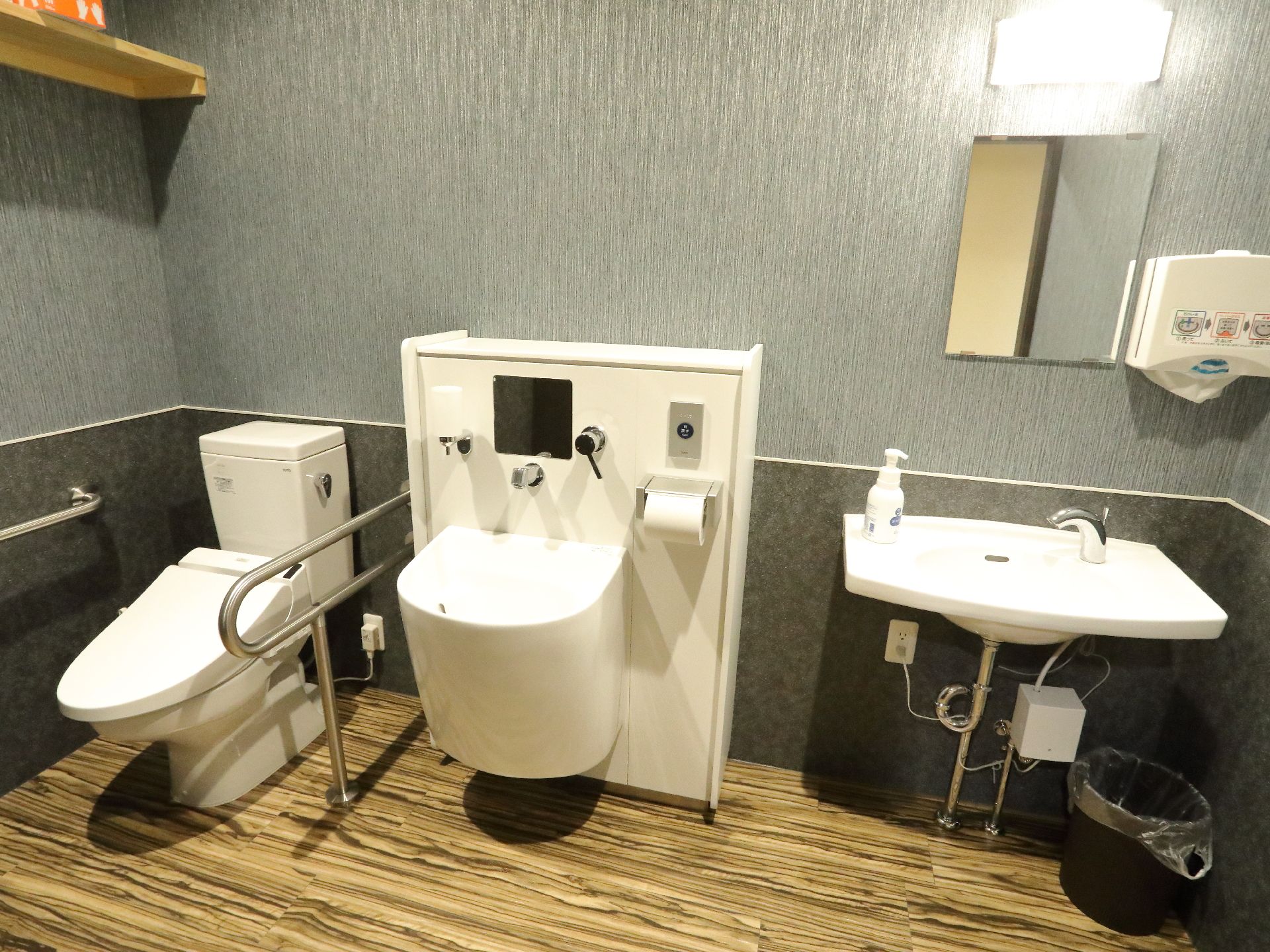 左側から順に便器があり、洗面台が2つ並んでいる。右側の洗面台には近くにペーパータオルがあり、その下側にはゴミ箱が置かれている。