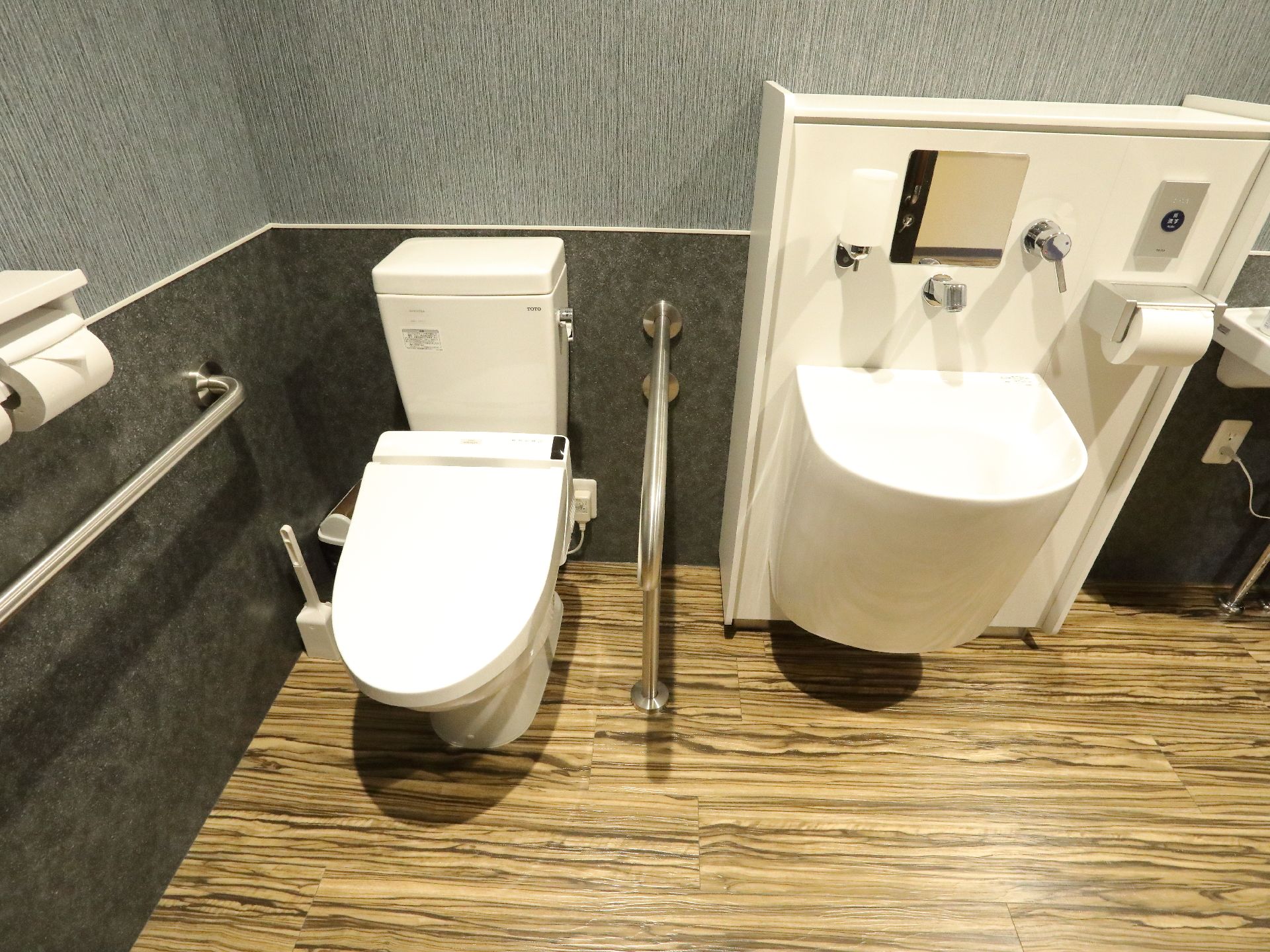 床は木目調になっている。左側には便器があり、その左右両側には手すりが取り付けられている。便器の左側にはトイレブラシが片付けられている。