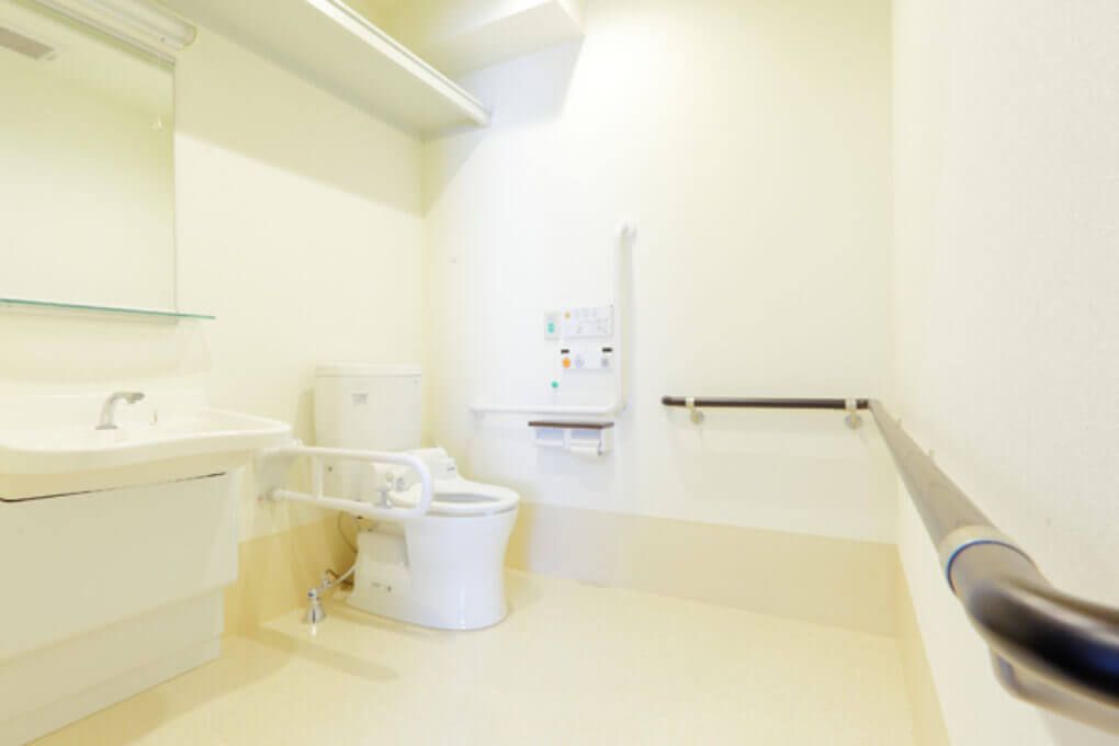 明るくスペースの広いトイレである。洗面台が完備されている。棚や手すり、呼び出しボタンが壁に設置されている。