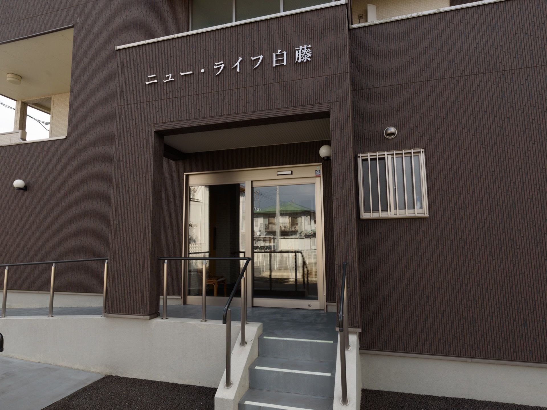 ダークブラウンの外壁に施設の名称が表示された屋根付きの入り口には階段の他、スロープが設置されている。