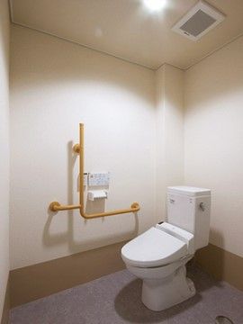 白い壁グレーの床の広いトイレには、便器の横にＬ型で木製の手すりとペーパーホルダーがあり、天井には換気口がある。