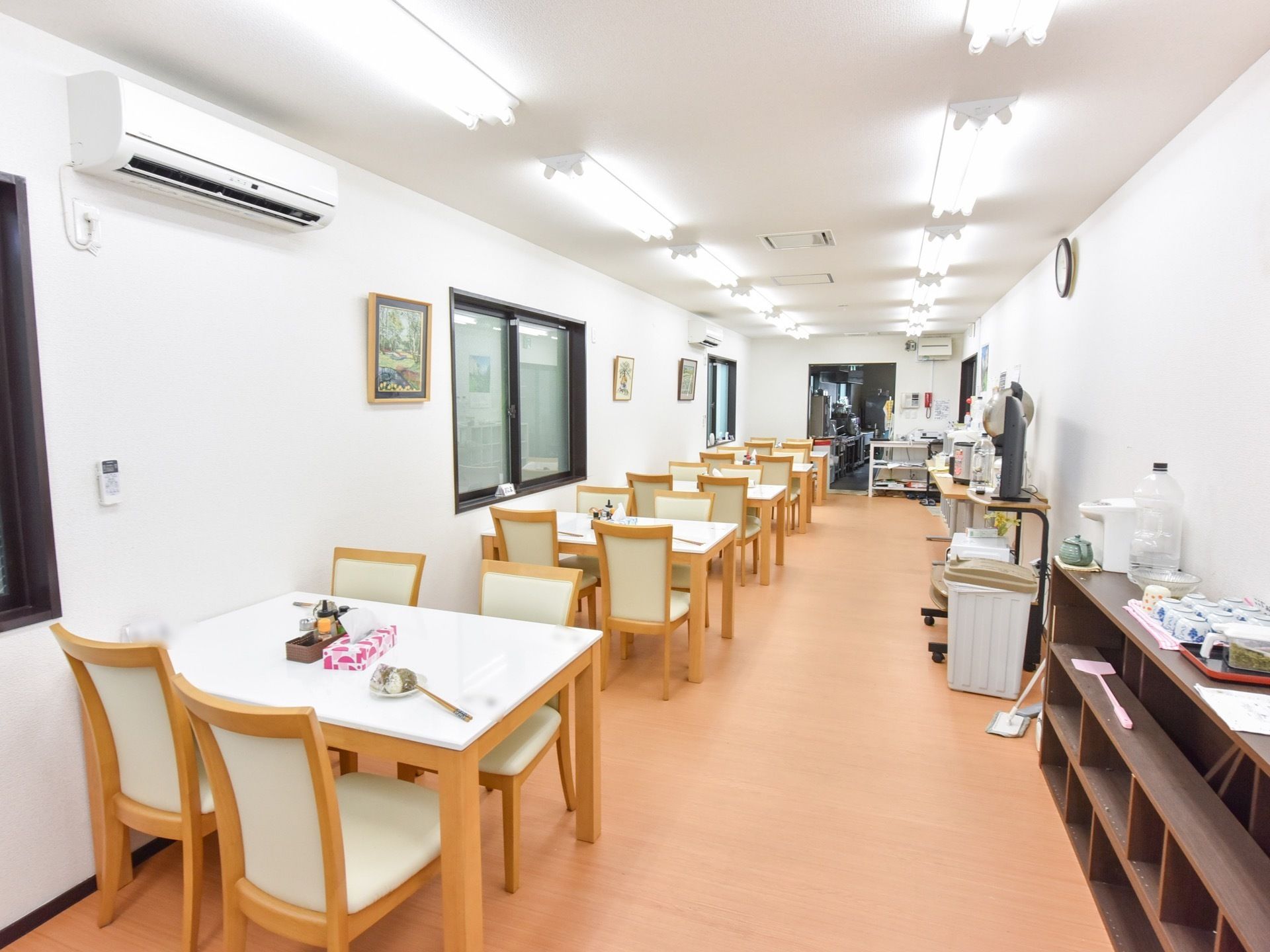 食堂には４人掛けのテーブルと椅子が複数並べられている。各テーブルには調味料やテッシュが置かれており、壁には窓、絵画や時計、エアコンがある。