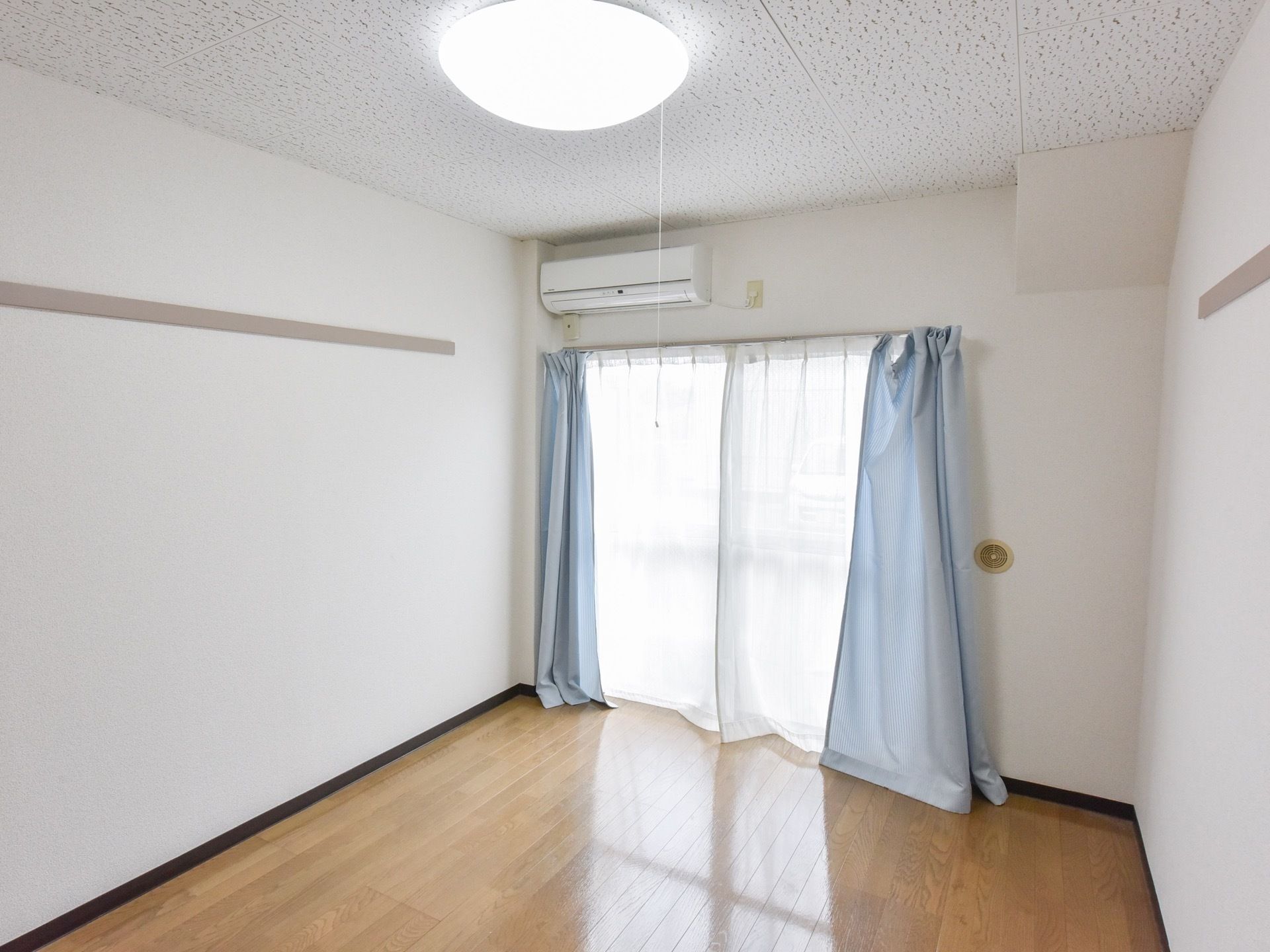居室は広々としており、窓にはレースと水色のカラーカーテンが掛けられている。壁にはエアコンが設置されている。床はフローリングである。