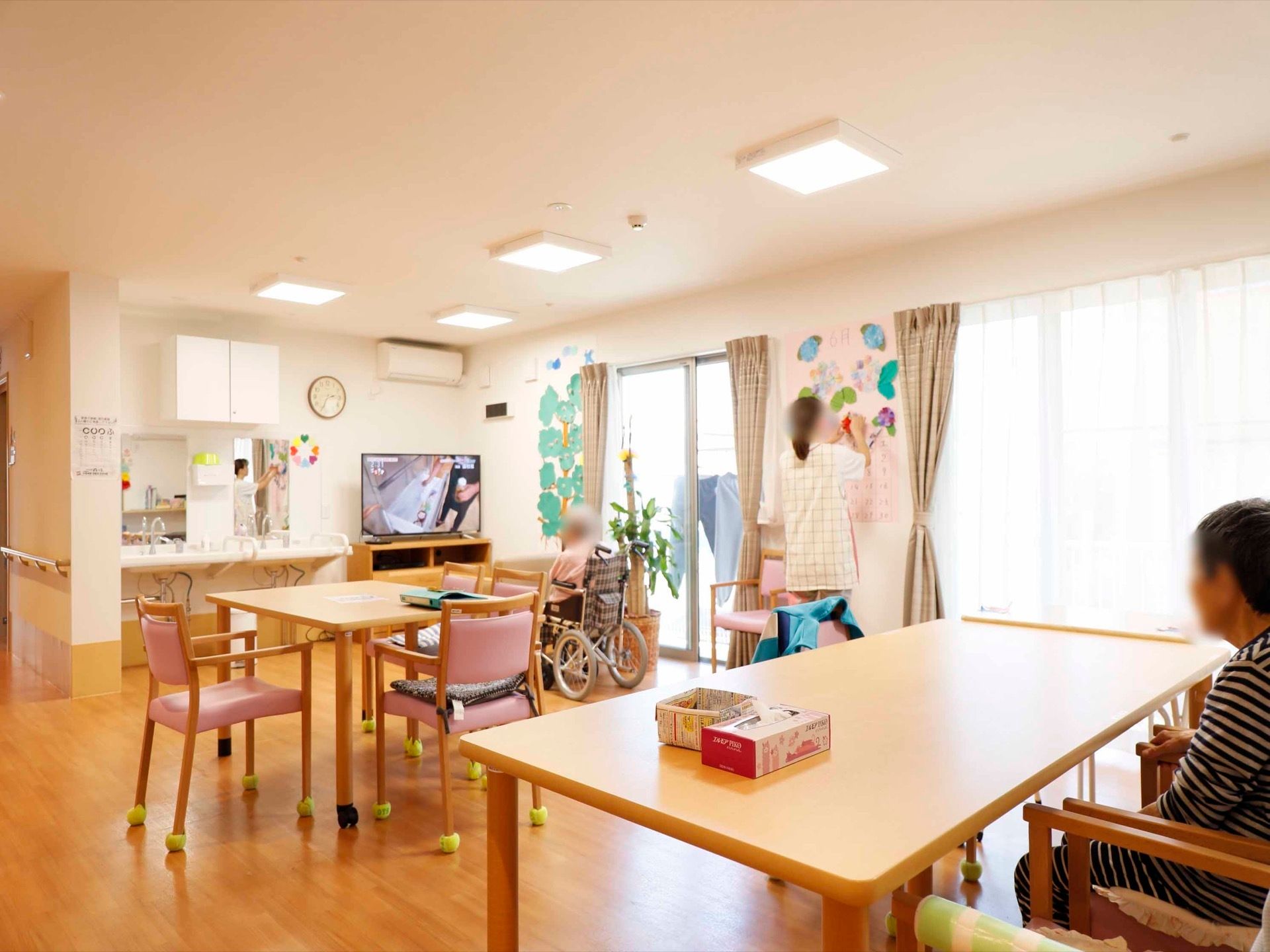 大きな窓が２つ設置された共有スペースである。テーブルと椅子が置かれ、入居者様がテレビを見ながら過ごしている。