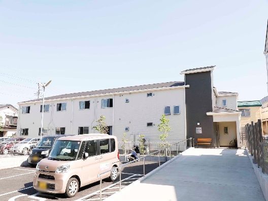 白い外壁の２階建ての建物は日当たり良好で建物の前には駐車場があり、車が止まっている。