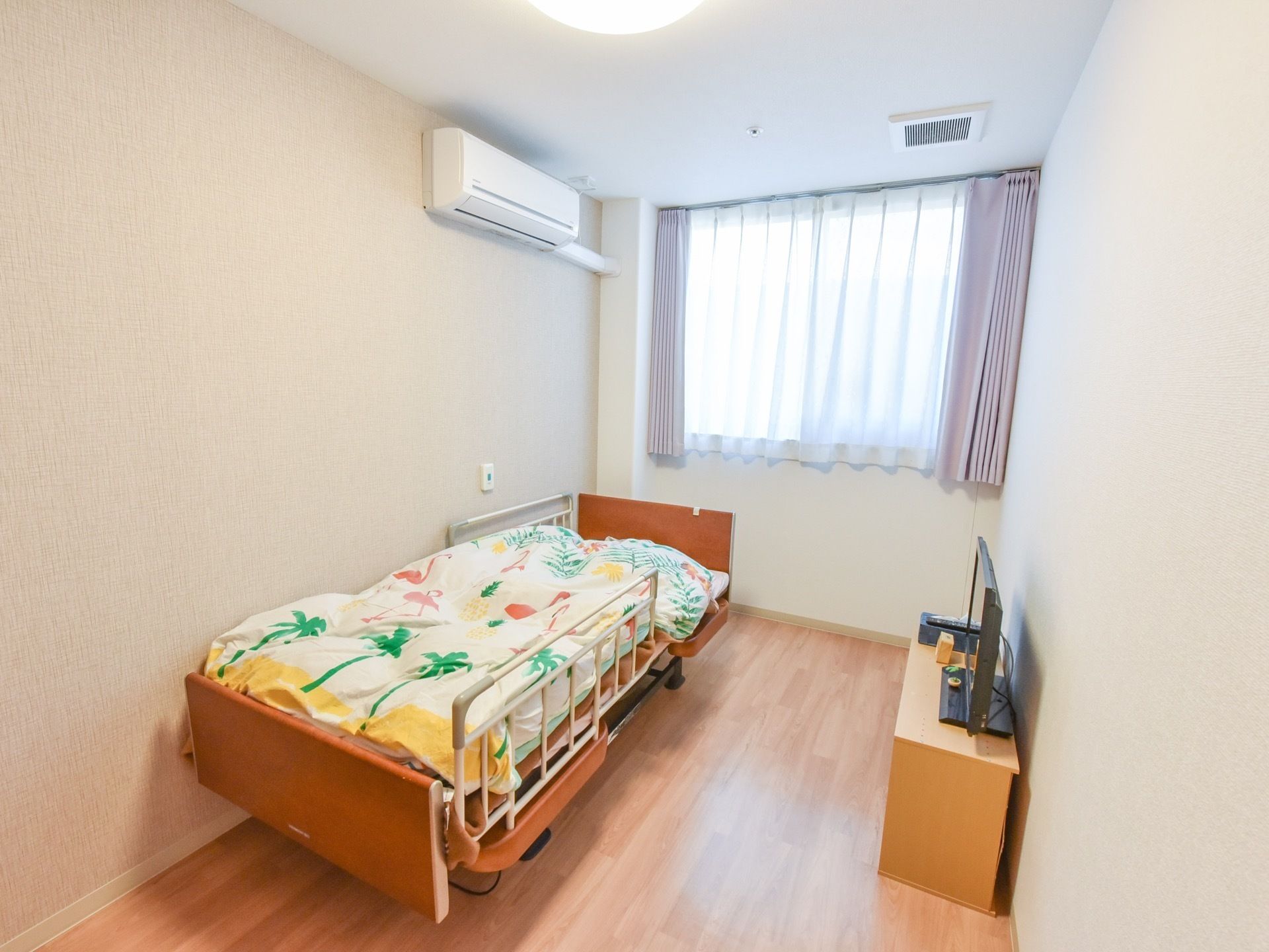 ゆったりとした居室には、正面に大きな窓があり、壁際に介護用ベッドを備えている。天井下にエアコンを取付けている。