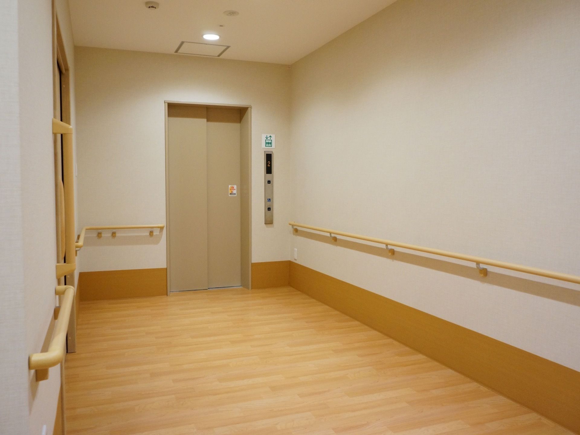 ベージュ色の扉のエレベーターが設置されている。目の前のフロアはフローリングで、壁には手すりが設置されている。