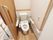 居室に備えているトイレスペースである。木目調の床に、白の壁際には、エル型の手すりを設置している。後方には、棚を設けている。