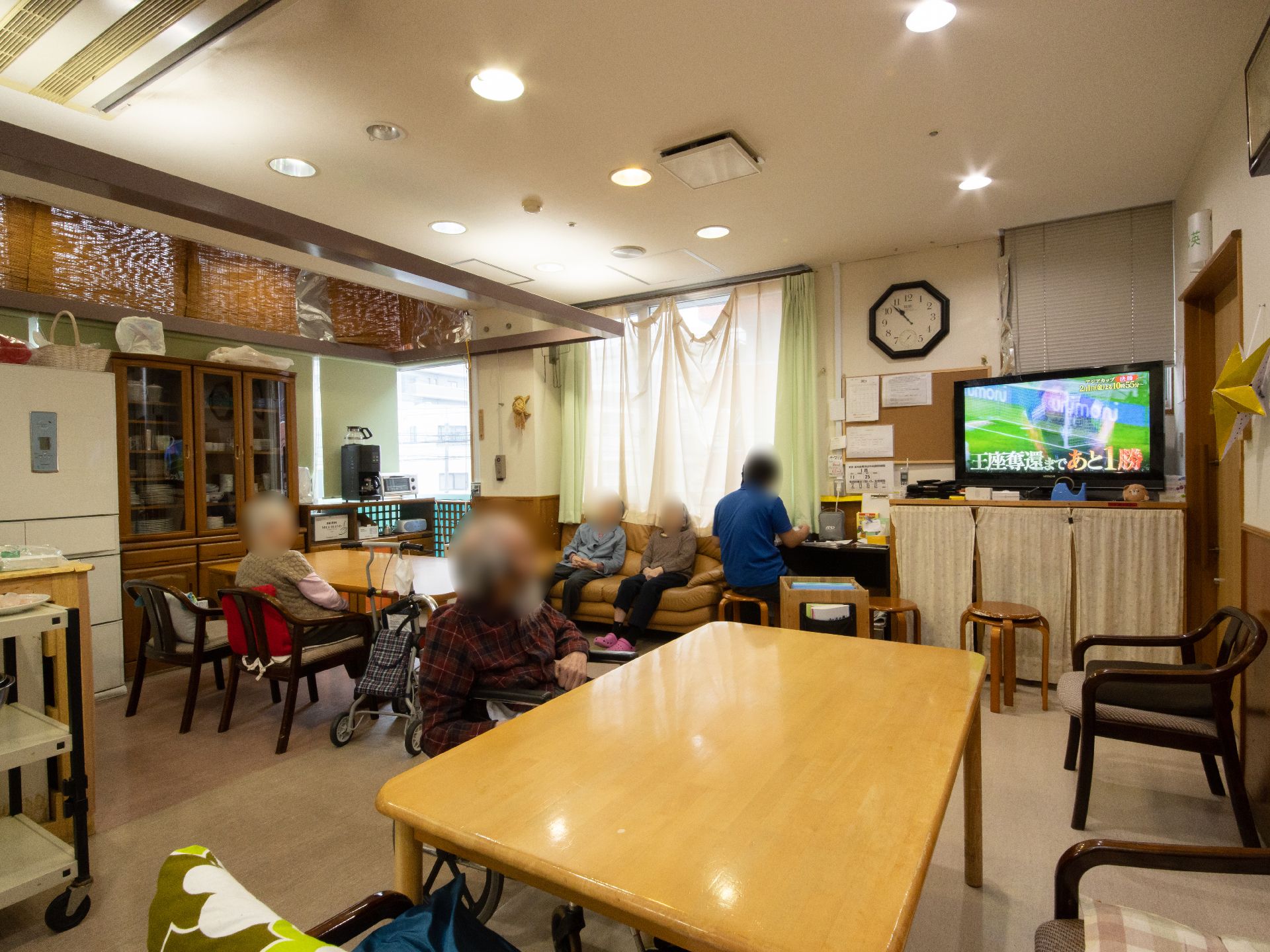 テーブルや椅子が配置され、ソファやテレビが置かれている共有スペースである。数名の入居者様と作業中のスタッフの姿が見える。