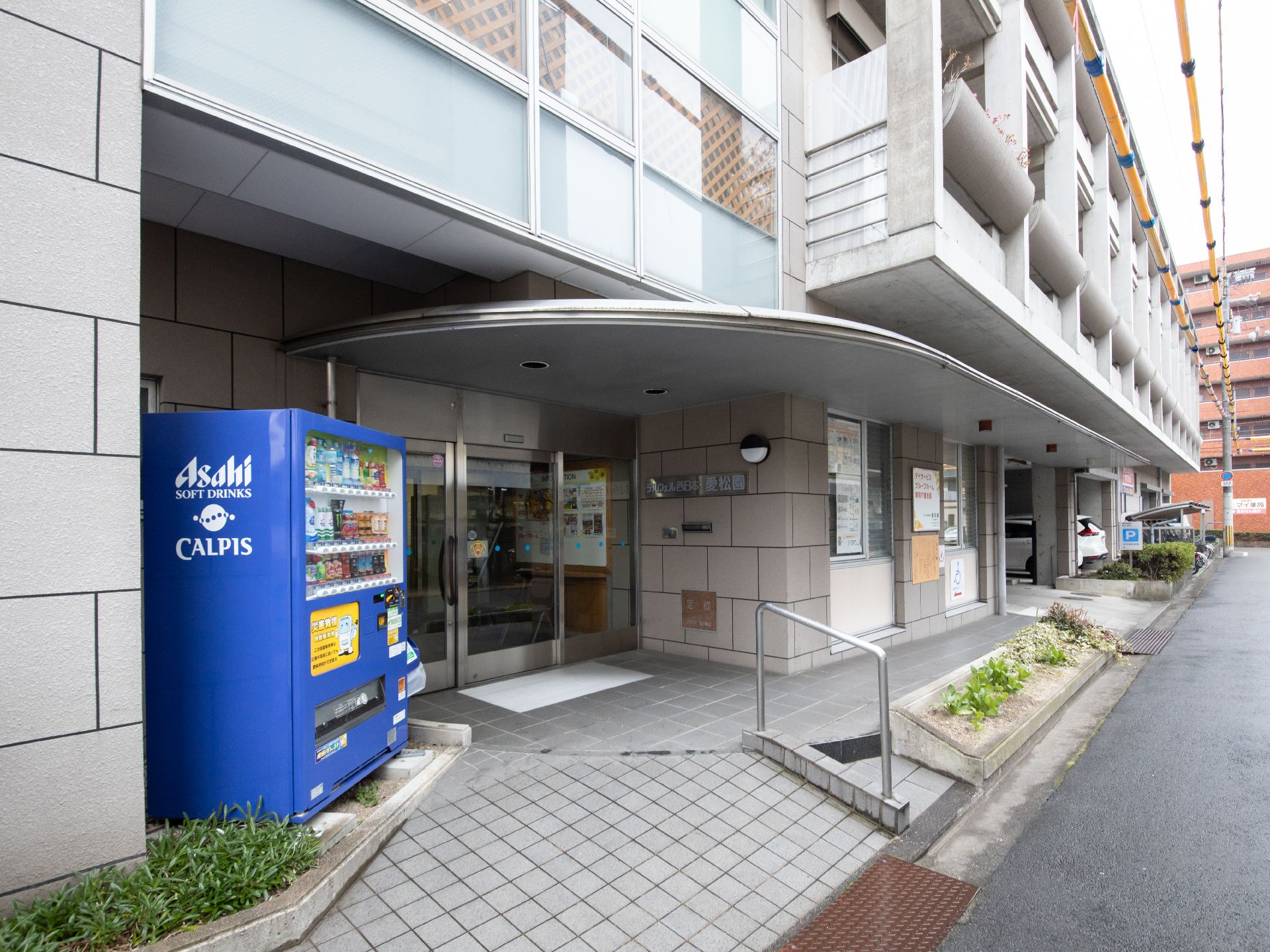 青い色の自動販売機が施設入口のそばに設置されている。道路から緩やかな傾斜で施設入口につながっている。