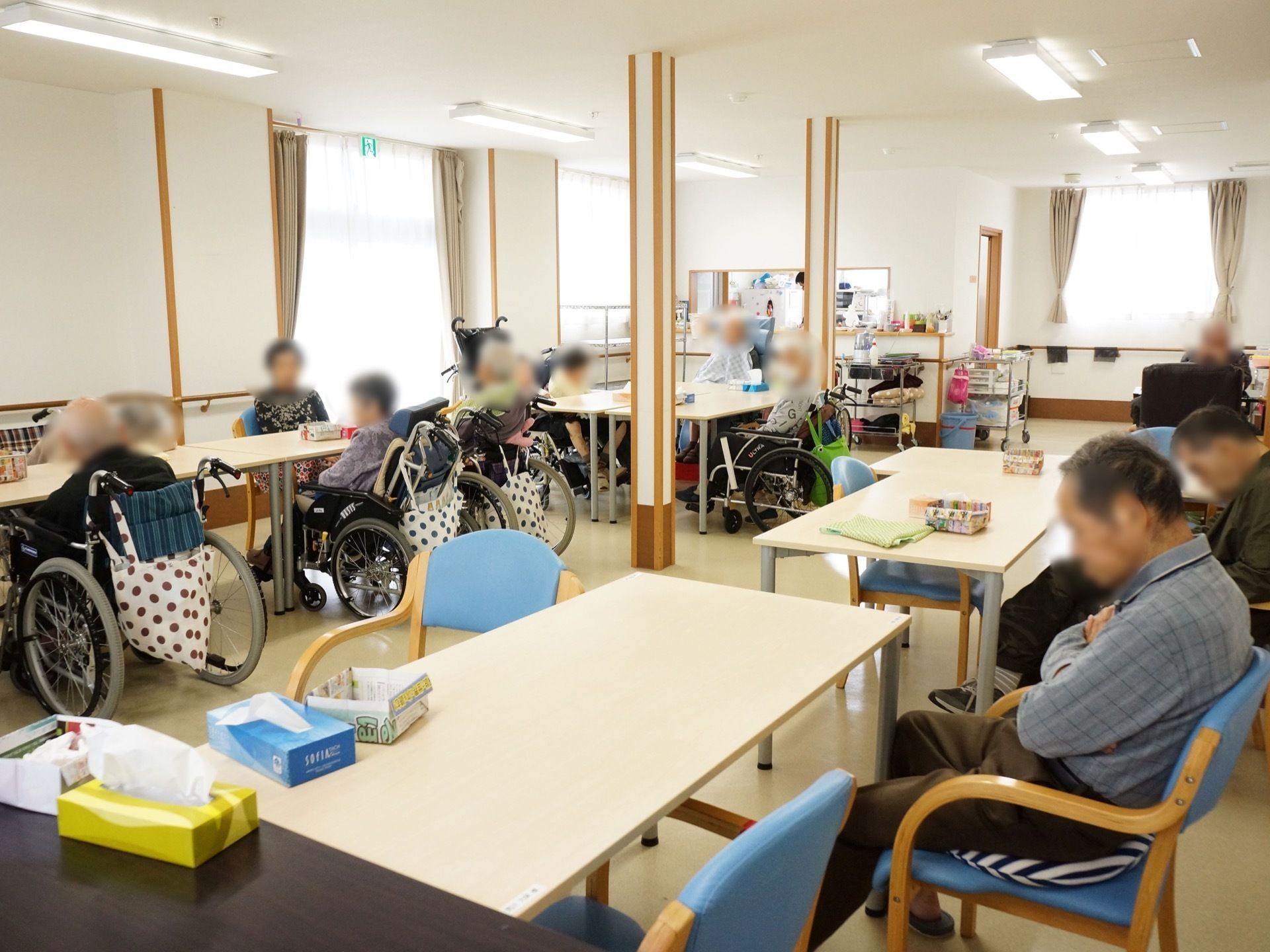 二列に並んでいるテーブルセットに、椅子や車椅子で座っている入居者たち