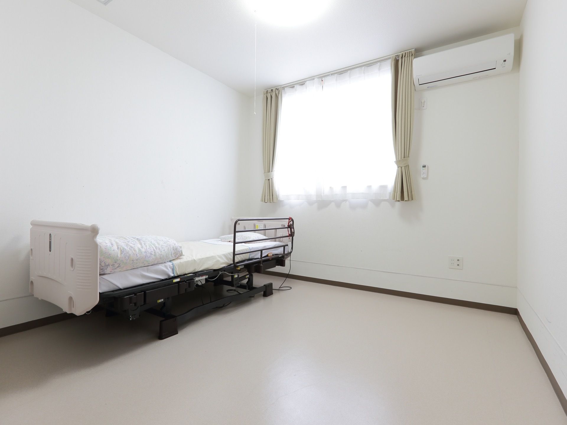 介護ベッドの設置されたシンプルな一人用の居室。壁には光が差し込む腰高の窓と、エアコンが設置されている。