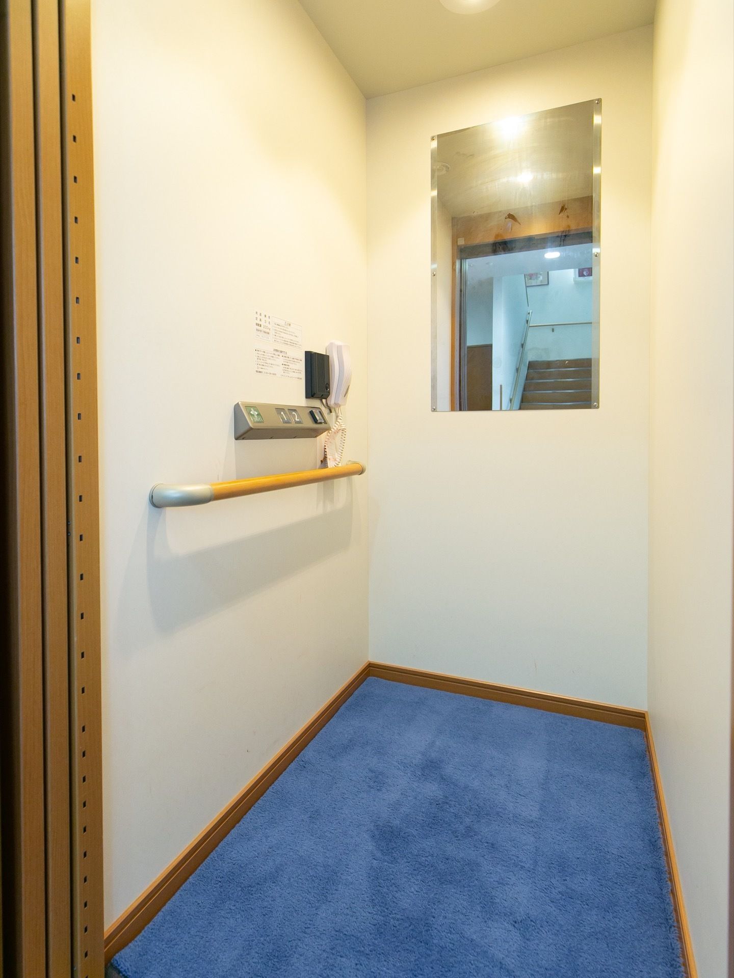 館内に備え付けられたエレベーターの図である。床には青い絨毯が敷かれている。奥の壁に大きな鏡がかかっている。