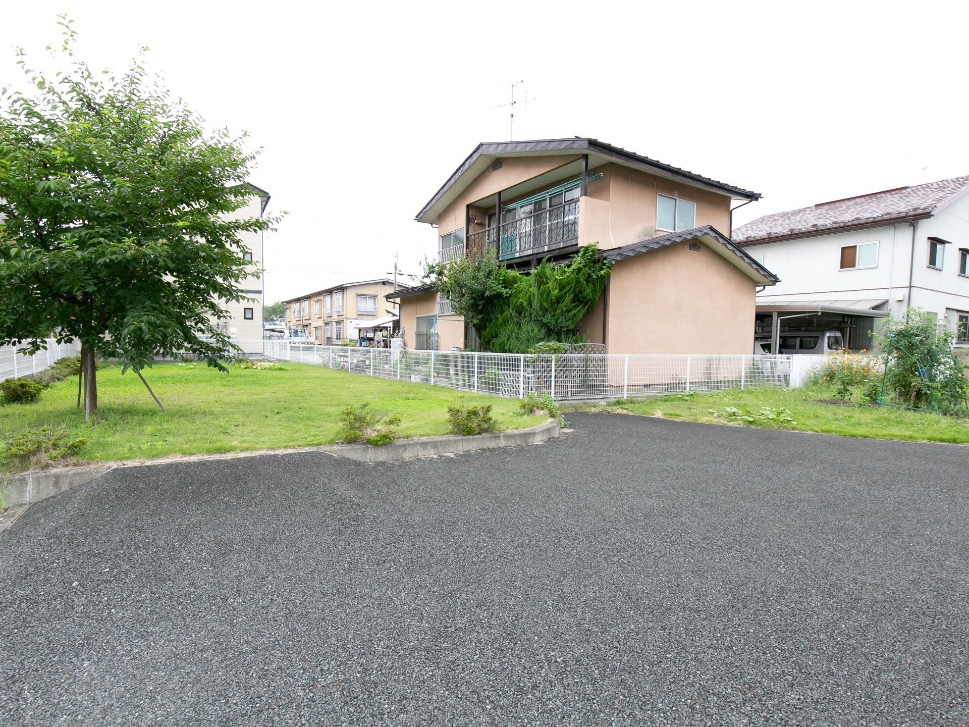 ピンク色の外壁の２階建ての建物が奥に建っている。白いフェンスで囲まれており、手前には舗装されたスペースが広がっている。