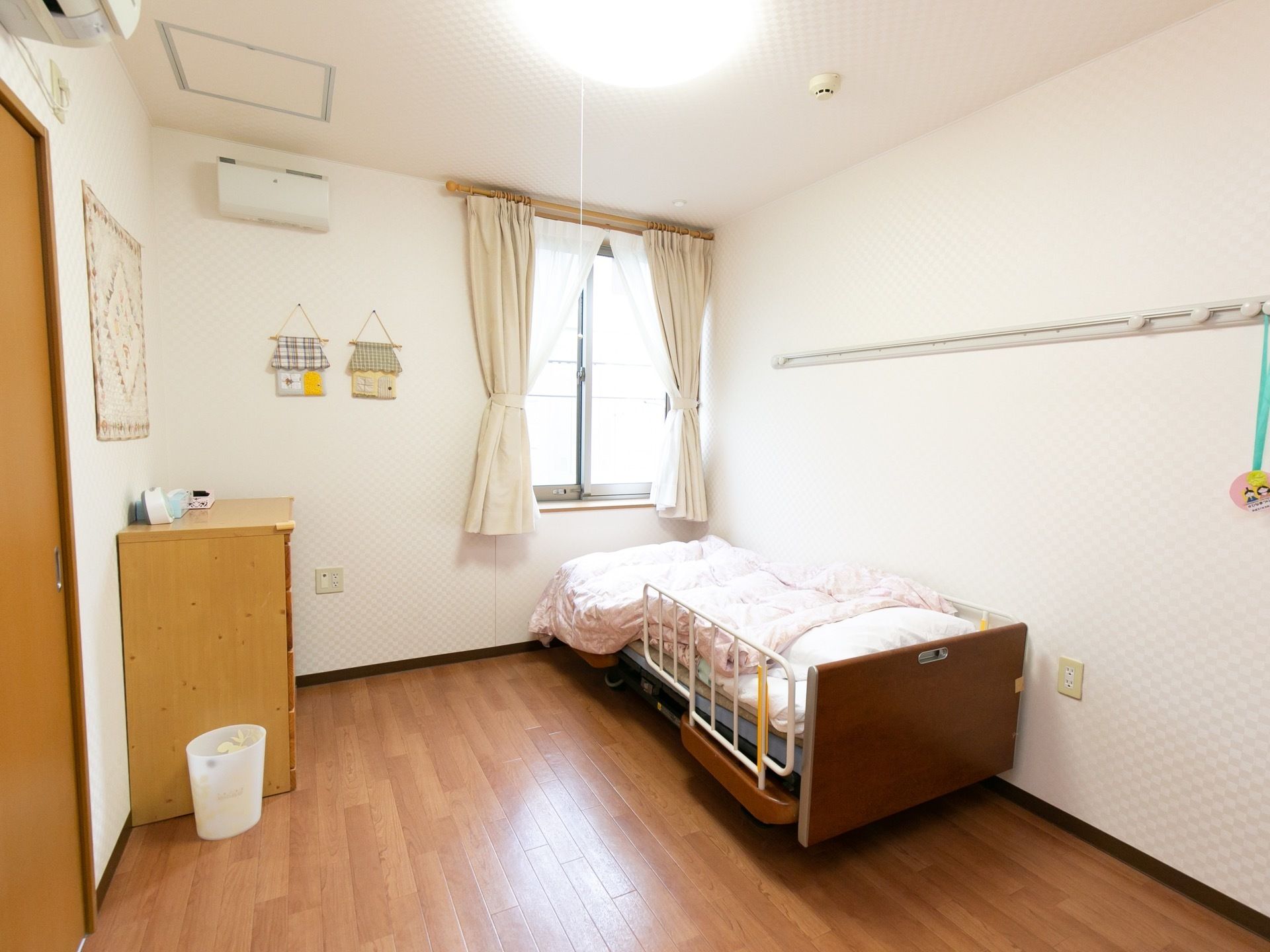 右奥に窓があり、その前に介護用ベッドがある。壁にはレールフックがある。チェストが１台ある。