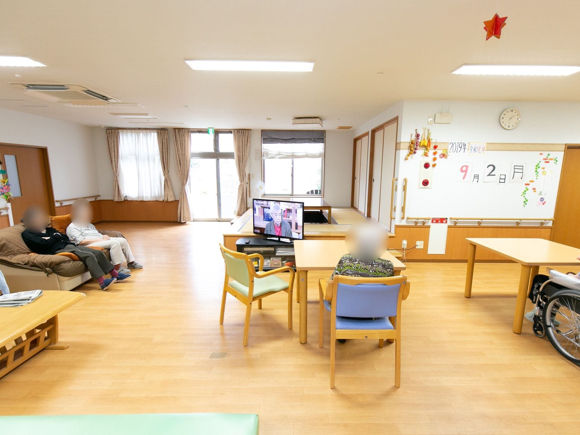 空間を広くとった共有スペースで、木製のテーブルがたくさん置かれている。あちこちで入居者様がくつろいでいる。