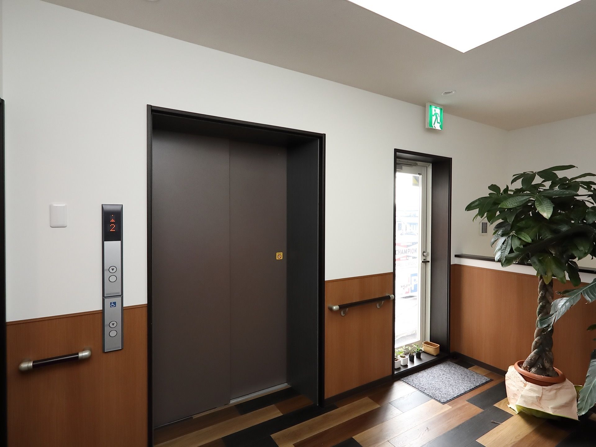 茶色の扉のエレベーターでドアの左側の壁に呼び出しボタンがある。右側には通用口があり、床に靴が並べられている。