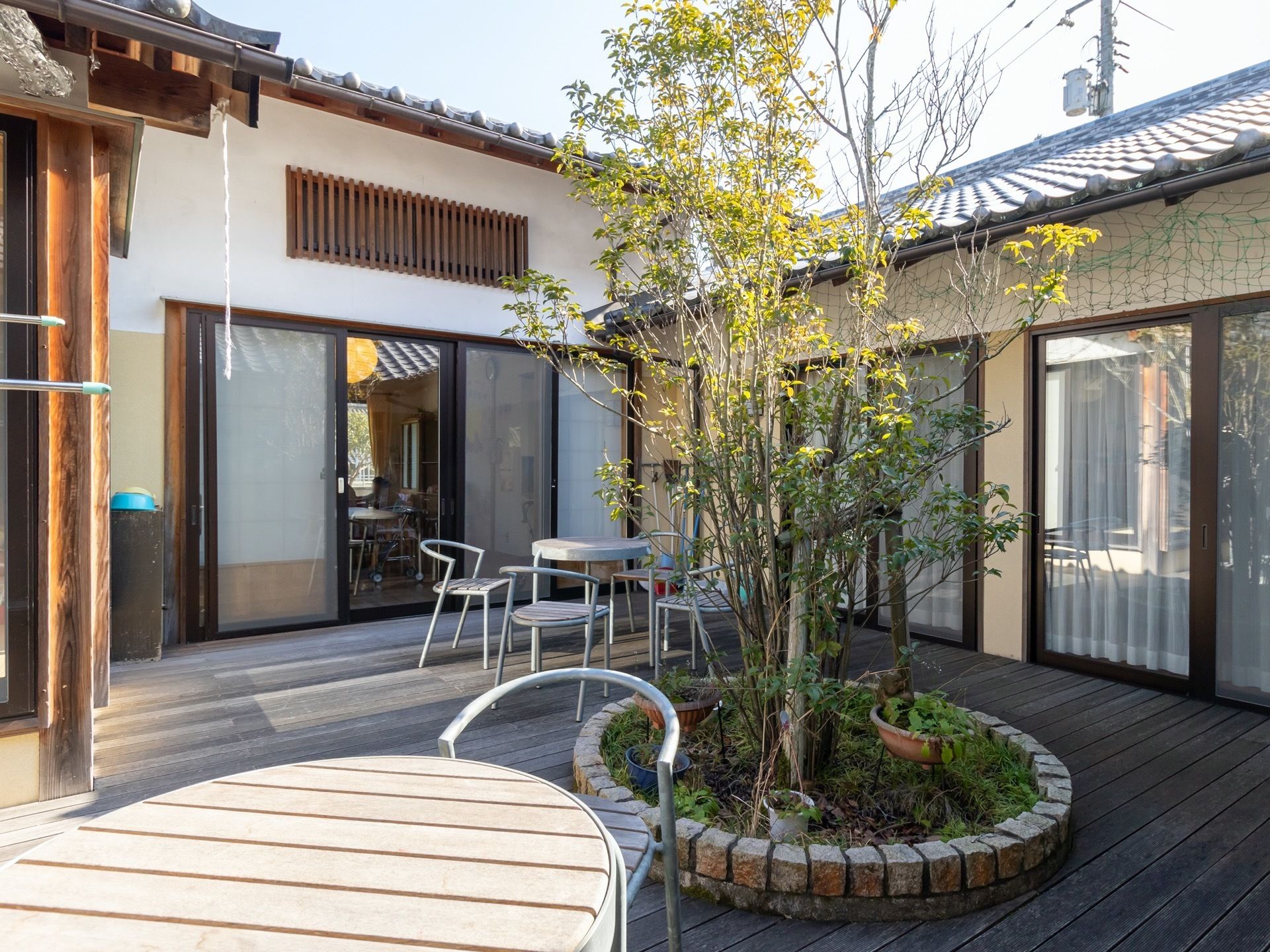 リビングから出入り可能なウッドデッキで、中央には木が植えられた花壇が設けられている。複数のテーブルセットが配置されている。