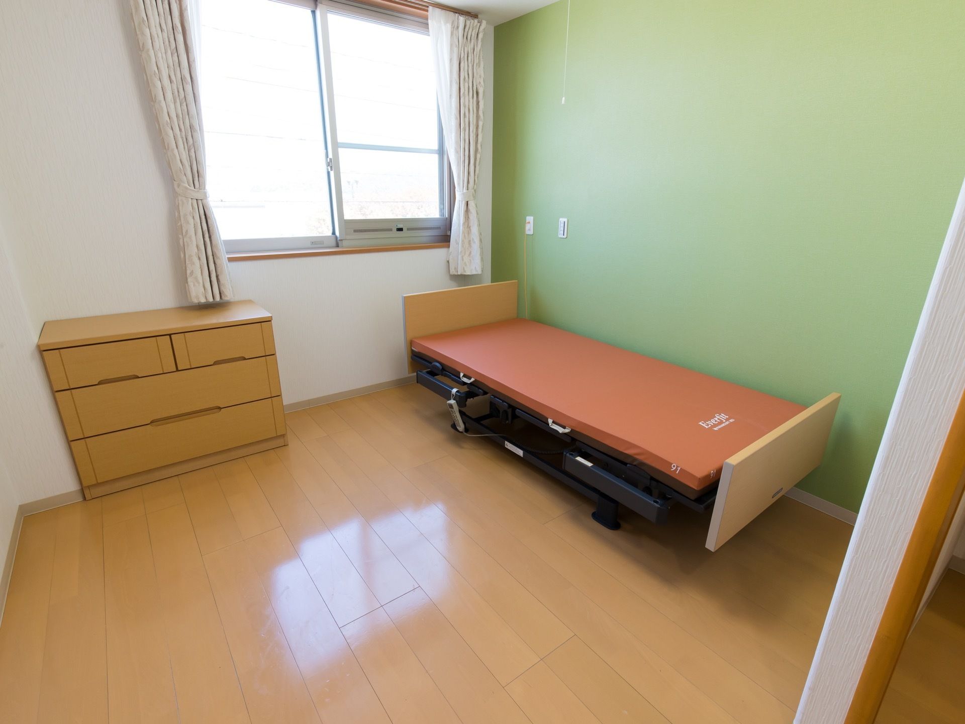 白と緑の２色の壁で構成された居室である。床はフローリングになっており、ベッドとチェストが置かれている。