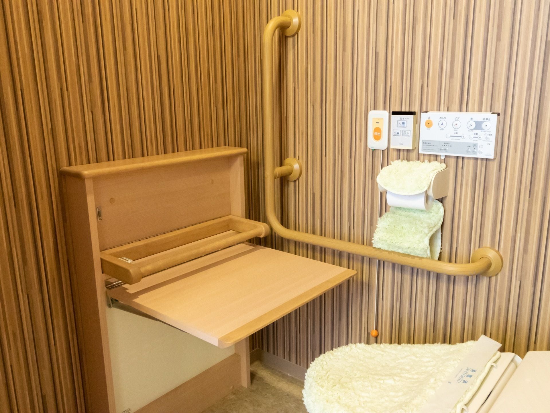 ストライプの壁紙が個性的なトイレは、天井が高く広々としている。自立支援のため、便器の前にテーブルのような器具が置かれている。