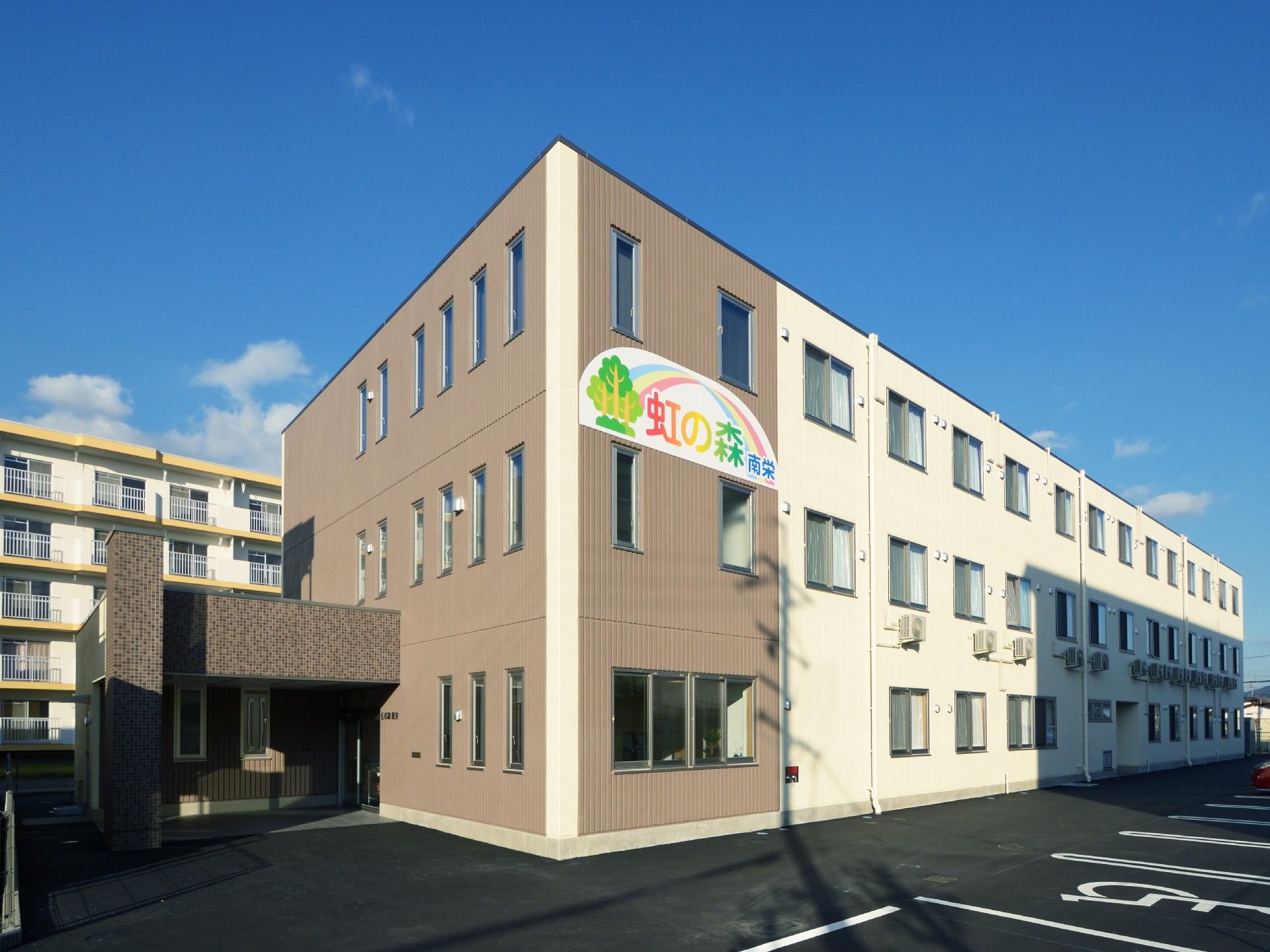 施設は３階建てで、２階と３階の間に施設名を表示した看板を掲げている。森と虹をモチーフとしたロゴは明るい印象である。