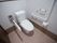 木目調の床と柄のある壁紙に囲まれたトイレである。温水洗浄便座があり、可動式とＬ字型の手すり、緊急ボタンが設置されている。